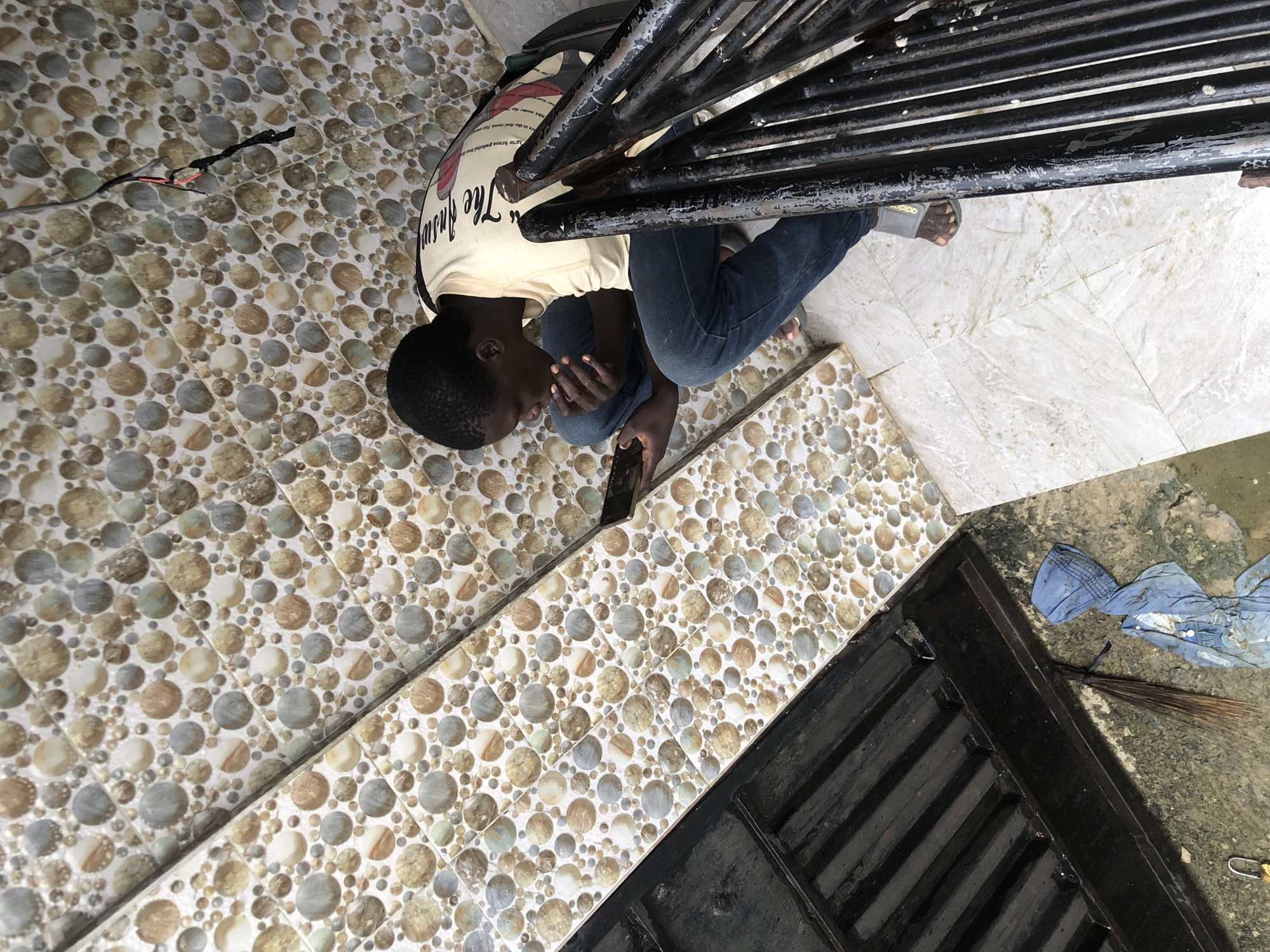 In Ado, Nigeria, a young person is crouched on a tiled staircase, engrossed in their mobile phone. They wear a light-colored t-shirt with text on the back and blue jeans, with grey sandals on their feet. The staircase is decorated with a mosaic of small, circular tiles in various earthy tones and shades of grey and white. To the left of the stairs, a dark, slatted metal grate covers a drainage area, with a broom and a bundle of blue cloth lying nearby on the ground. A black metal railing runs alongside the stairs. The scene appears to be an outdoor or semi-outdoor setting, possibly a residential building or a public space.