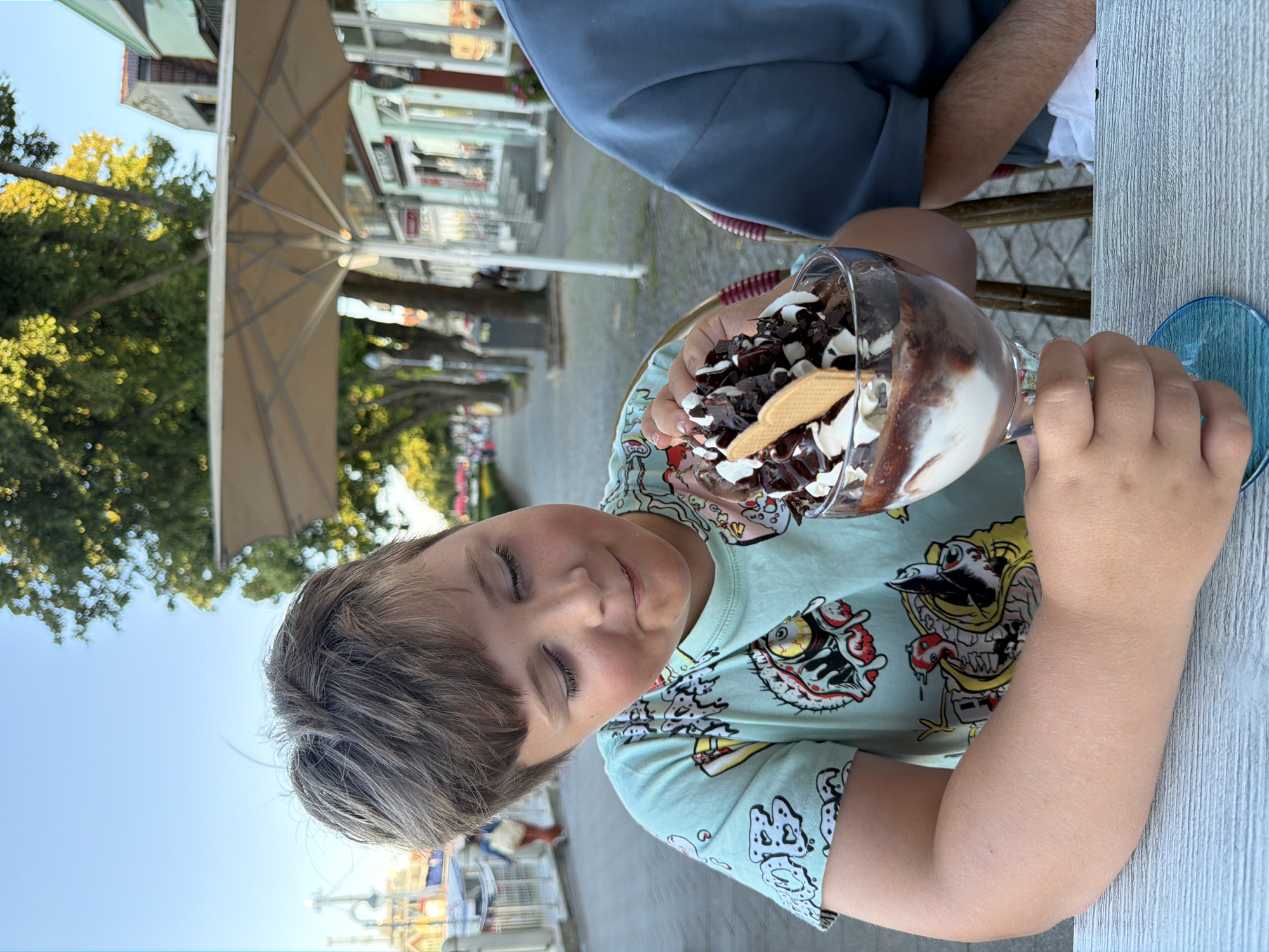 In Rostock, Germany, a young boy is seated at an outdoor cafe table, holding a large glass of ice cream. The ice cream is topped with whipped cream, chocolate sauce, and chocolate shavings. The boy wears a light blue t-shirt with a colorful, cartoonish design featuring monstrous figures. He is looking down at the ice cream with a slight smile. Beside him, a person wearing a blue shirt is also seated at the table. The background shows a treelined street with buildings, suggesting an urban setting, possibly a promenade or waterfront area given the presence of what appears to be a dock or harbor in the distance. An outdoor umbrella is visible behind the boy, providing shade. The sky is clear and blue.