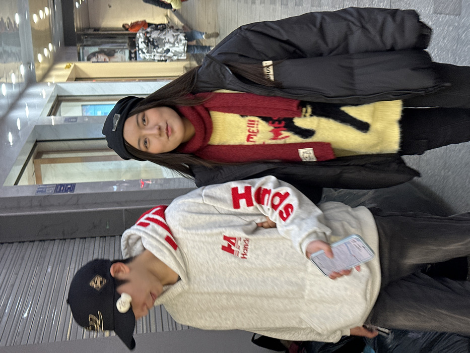Two young East Asian individuals are depicted standing on an urban commercial street in Jiangjin, China. The individual on the left is a male-presenting subject wearing a black baseball cap with a white circular emblem on the brim, a light grey hooded sweatshirt with red "Honda" branding on the chest and vertical red text on the right sleeve, and dark trousers. He is looking downwards and to his left, holding a light-colored smartphone in his right hand. The individual on the right is a female-presenting subject, wearing a black beanie, a red knitted scarf, a yellow fuzzy sweater with a black graphic and red "me!!!" text, a black puffer jacket, and dark trousers. She is facing the camera with a slight smile. The subjects are in close proximity, positioned side-by-side. The background features storefronts with glass windows and interior lighting, displaying blurred merchandise and signs with Chinese characters. A paved pedestrian walkway is beneath the subjects. Blurred figures of other pedestrians are visible further down the street.