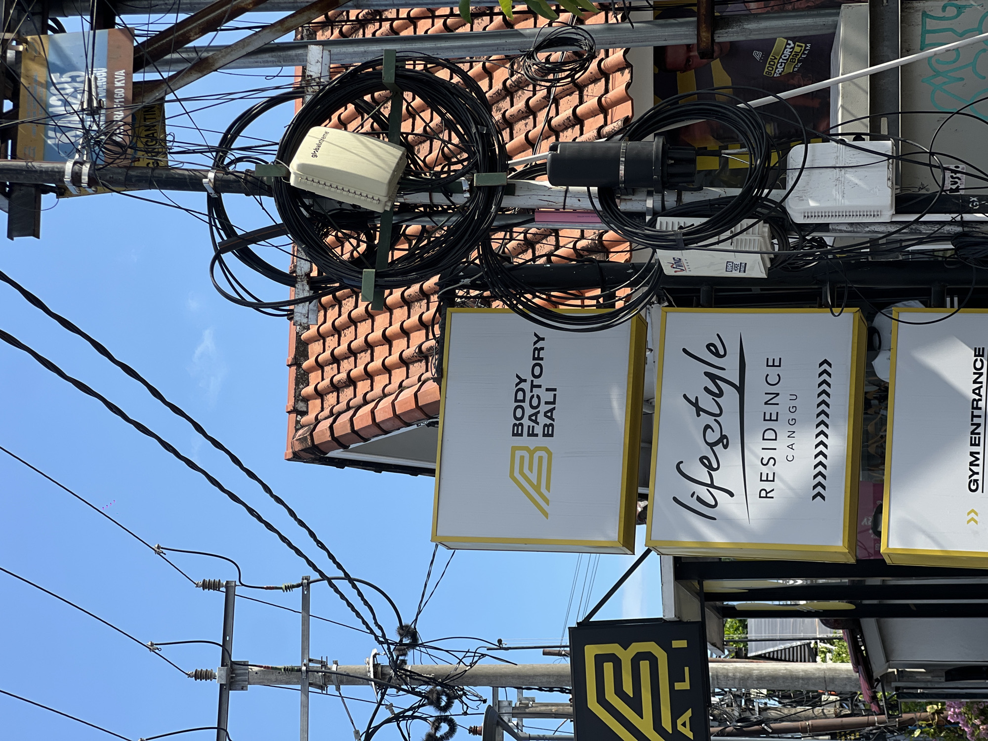 The image shows a chaotic overhead tangle of black wires and utility equipment mounted on a metal pole against a bright blue sky. The pole is near a building with orange tile roofing. Three signs are visible, advertising "BODY FACTORY BALI," "lifestyle RESIDENCE CANGGU," and "GYM ENTRANCE." Additional signage is partially visible below. According to text captured from the image, this location is in North Kuta, Indonesia. Utility equipment visible includes boxes from globalxtreme and VING. Other text captured includes "SS RAP160 KVA" and "GANGAN TIN."