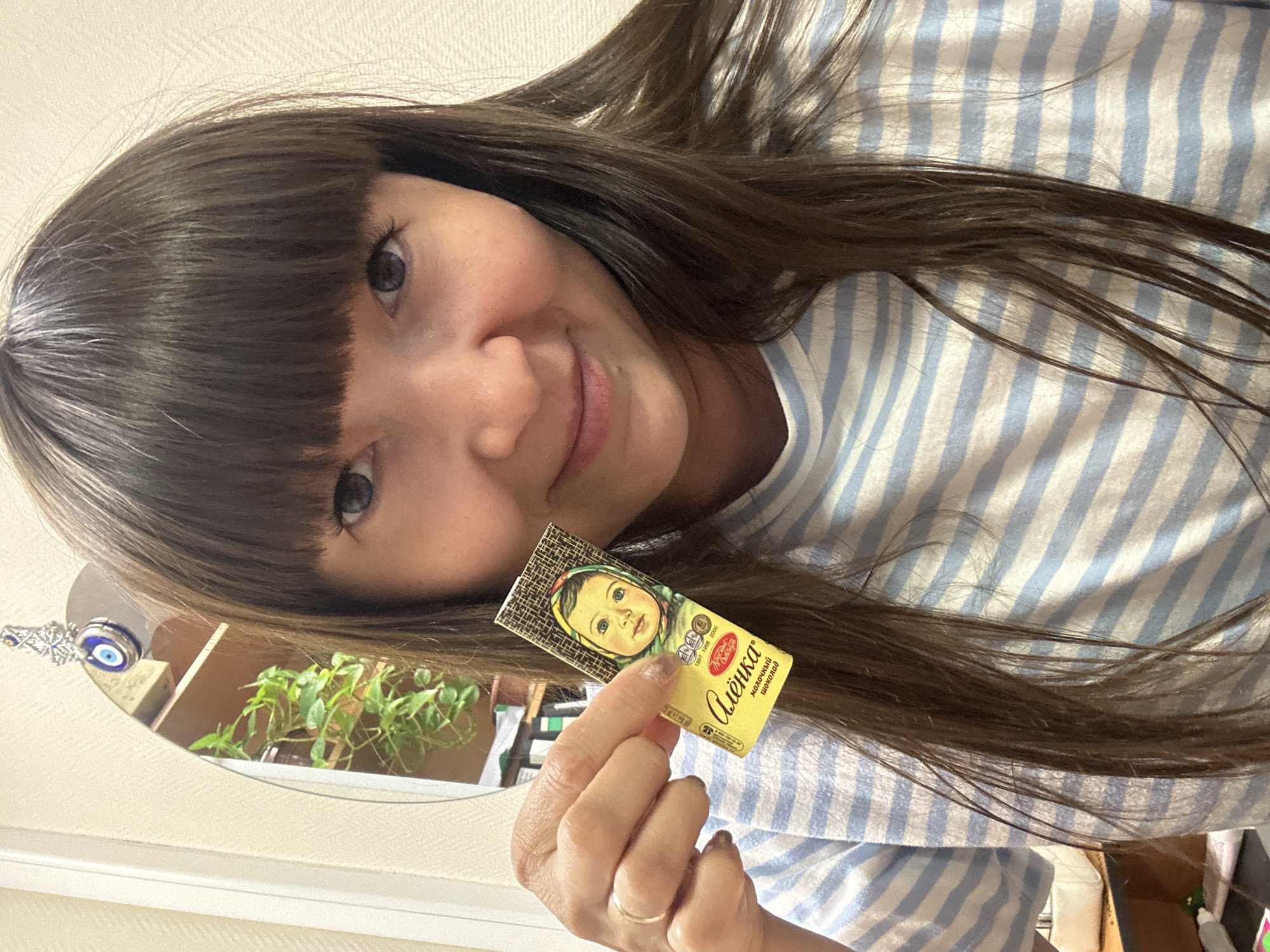 A young woman is captured in a close-up, slightly tilted selfie, holding a small chocolate bar. She has long, straight dark brown hair with a full fringe, and is smiling gently at the camera. Her eyes appear light-colored, possibly blue or gray. She is wearing a light blue and white striped top.
In her right hand, she holds a yellow-packaged chocolate bar. The packaging features a classic image of a baby girl in a headscarf, and prominent Cyrillic text identifies it as "Алёнка" (Alyonka) milk chocolate ("молочный шоколад"). Other visible text includes "ГОСТ" (GOST, a quality standard) and a date range, possibly "1965 2005". A simple gold ring is visible on her right ring finger, and some fingernails have red polish.
The background, partially visible as a reflection in a mirror, shows a light-colored wall, a wooden shelf, and a potted green plant. An "evil eye" charm, featuring a blue eye design on a silver-colored key or ornament, hangs near the mirror. The overall setting appears to be an indoor space, likely a home, suggesting a casual and personal moment. The time of day appears to be daytime due to the natural lighting. The woman seems pleasant and content.