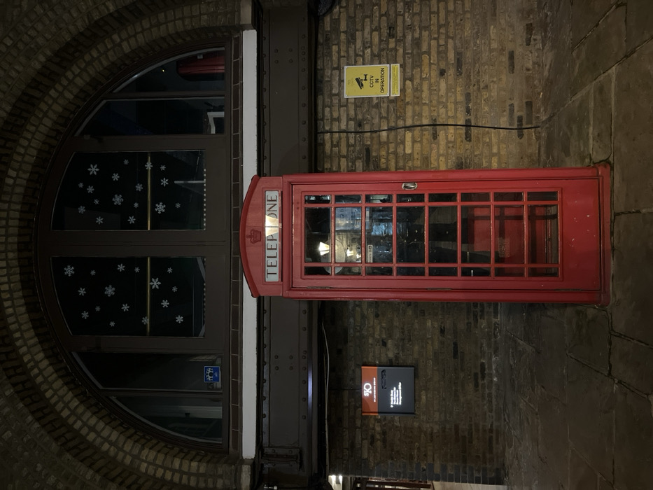 A red telephone booth stands next to a brick building with a large, arched window decorated with white snowflakes. Adjacent to the booth, a sign reads "TELEPHONE" in bold, white letters. Above, a "CCTV in Operation" sign is affixed to the brick wall. Below, a smaller, rectangular sign displays text and a graphic. The surrounding area is constructed of brick and stone, suggesting an urban, possibly historical, setting.