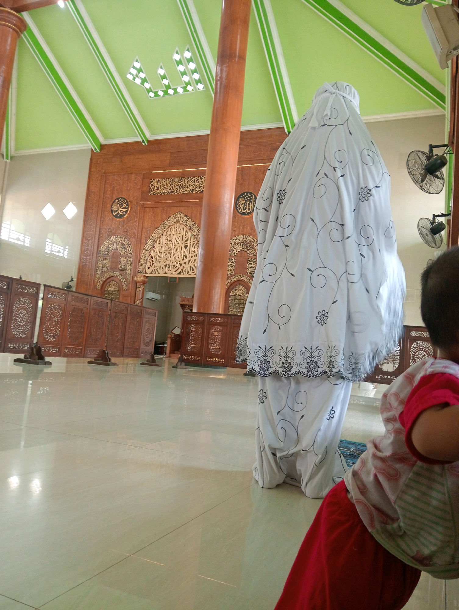 In a room with a light green ceiling and wooden pillars, a person in a white prayer dress with black floral patterns is standing. The person's face is not visible. The prayer dress is long and covers the entire body, including the head. To the right of the standing person, a young child is seen from behind, wearing a pink and white striped shirt and red pants. The child's head is turned to the right, and their face is not visible. The room has polished light-colored floors and wooden screens with intricate carvings. In the background, there is a large wooden wall with Arabic calligraphy and decorative carvings. Two electric fans are mounted on the wall. The scene appears to be indoors, possibly in a mosque in Soko, Indonesia.