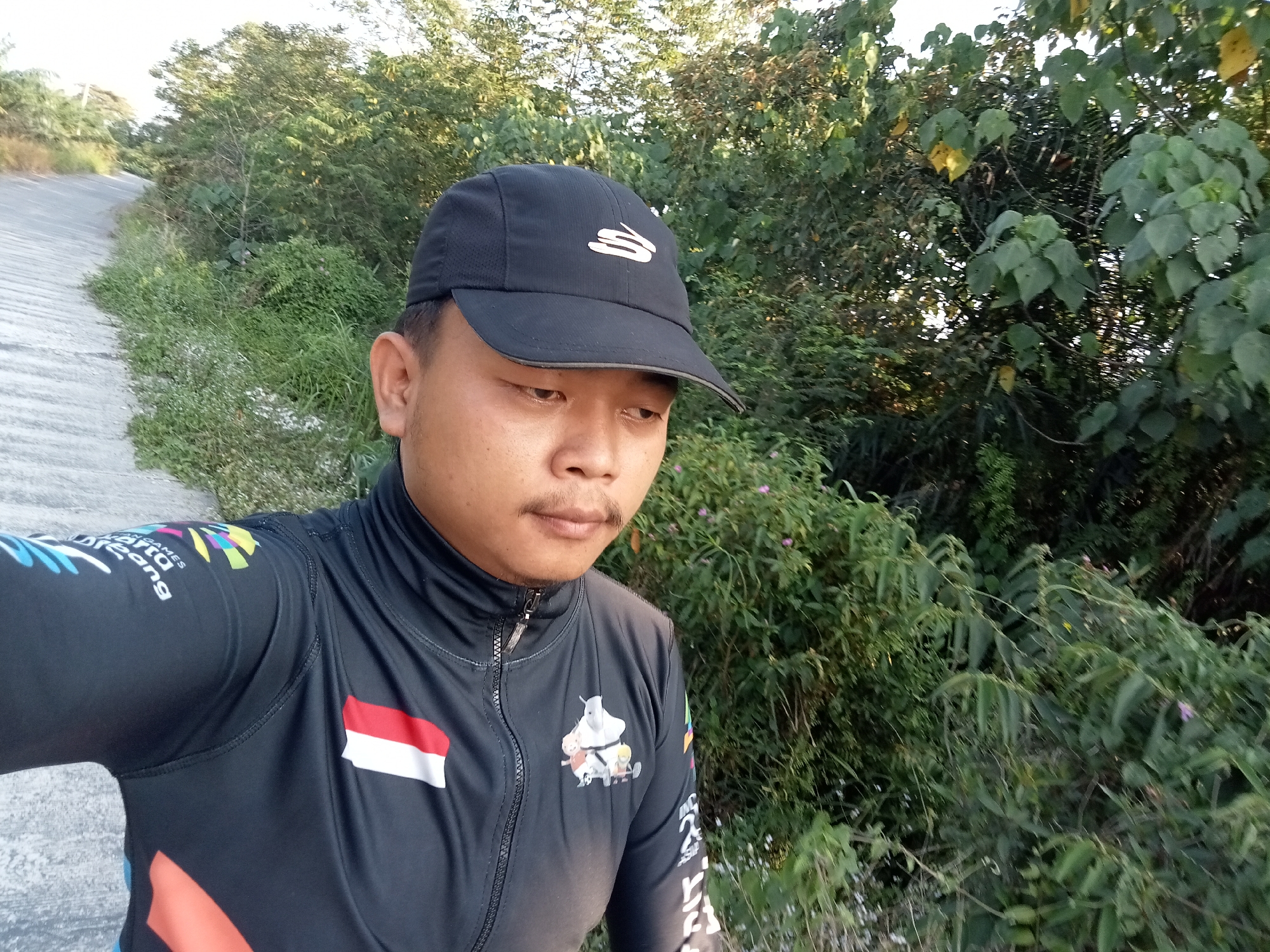 A medium shot captures a man wearing a black baseball cap and a black zip-up jacket with colorful graphics and an Indonesian flag patch on the left chest. He is standing outdoors with dense green foliage behind him and a concrete pathway to his left. The man's face is turned slightly down and to the right, with his mouth open. The overall lighting suggests it is daytime, possibly late afternoon, given the warm tones. The environment appears to be a rural or semi-rural area in Teluk Kenidai, Indonesia, characterized by natural vegetation and a paved path.