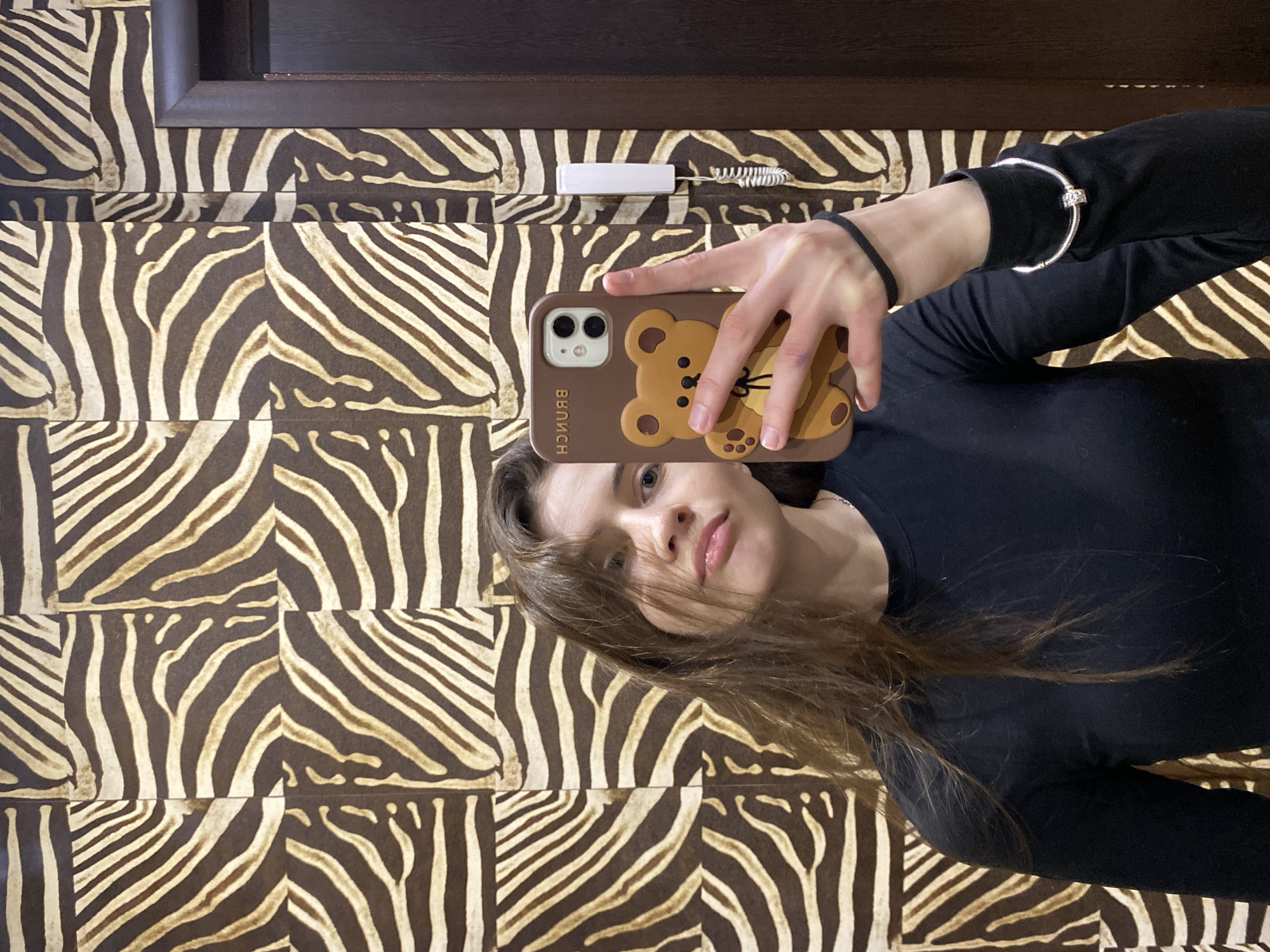 This mirror selfie shows a young woman, likely taken indoors in Votkinsk, Russia. The original image is rotated 90 degrees clockwise. The woman, with light brown hair falling over her face, wears a black long-sleeved top and gazes directly into the camera with a neutral expression. She holds a smartphone, likely an iPhone given its camera setup, encased in a brown silicone cover featuring a cartoon teddy bear design. The word "BRUNCH" is visibly printed vertically on the side of the phone case.
On her right wrist, she wears a silver-toned charm bracelet, while her left wrist, holding the phone, has a black hair tie or band. The background is dominated by a distinctive wall covering, appearing to be square tiles or wallpaper with a dark brown and light beige zebra-print pattern. Above the woman, a white intercom unit with a coiled cord is mounted on the wall, and a dark wooden frame, possibly part of a door or mirror, is visible at the top edge of the image. The setting suggests an indoor space, possibly a hallway or entrance area.