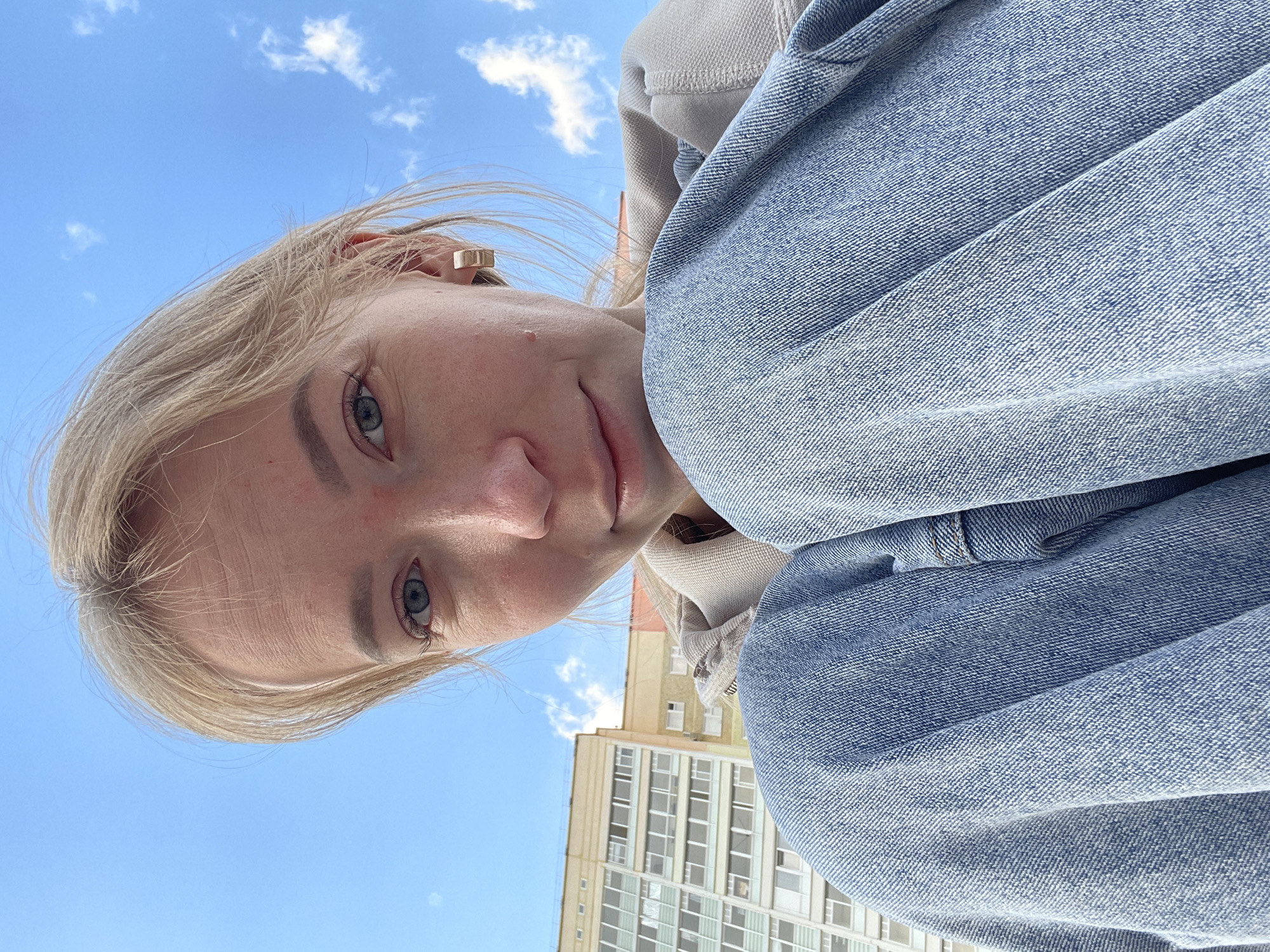 A low-angle selfie taken in Perm, Russia, features a young woman with fair skin and blonde hair. Her eyes are blue, and she is wearing a square earring. She is crouched down, knees pulled to her chest, and is wearing light-wash denim jeans. A light-colored hoodie is visible beneath the denim. Behind her is a light blue sky with scattered white clouds. In the background is a tall, light yellow building with multiple windows.