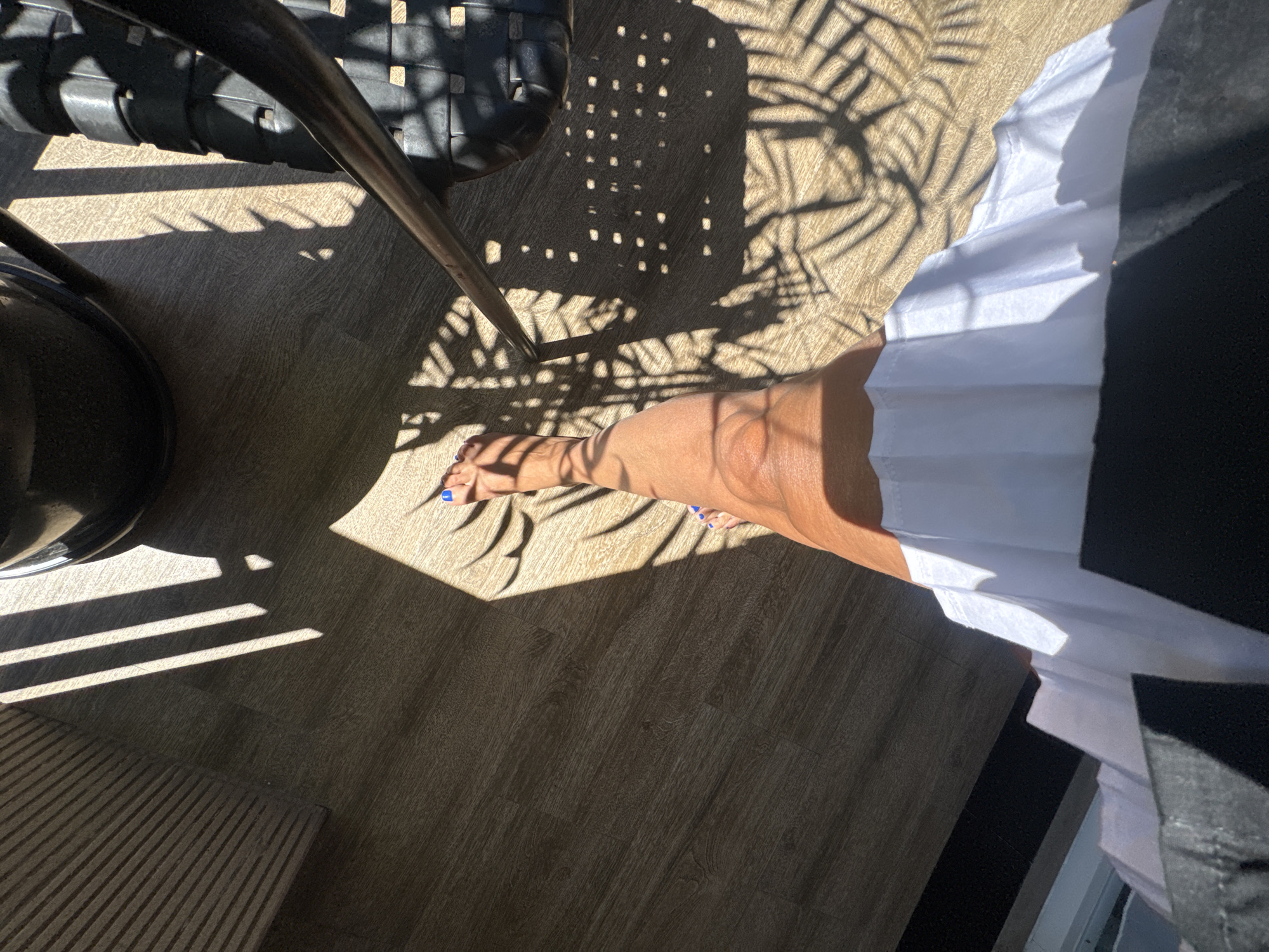 Here's a concise, forensic description of the image:
The photograph, taken from an elevated perspective, shows a partial view of an interior space with dappled sunlight and shadow patterns suggestive of a palm tree outside. The floor is wood-toned. A portion of a black, woven chair is visible, along with its metal leg. A person's legs and feet are present in the scene; toes are painted with blue nail polish. The person is wearing a white garment with dark material. Based on the presence of the tropical shadow patterns and general aesthetic, it is plausible that the location is Kuta, Indonesia.