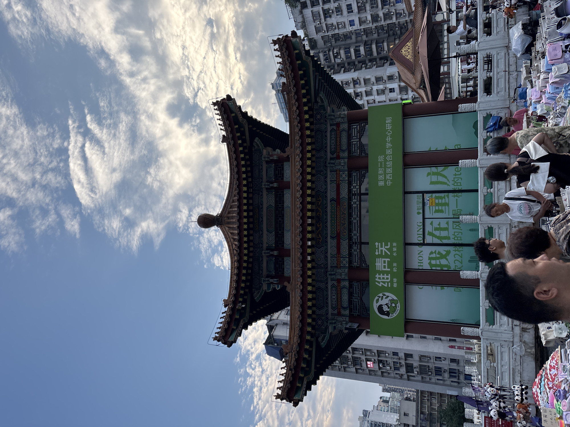 The image depicts a bustling street scene in Yuzhong District, China, dominated by a traditional Chinese architectural structure adorned with intricate carvings and a multi-tiered, ornate roof. A large green banner on the building features Chinese characters and a logo of a woman with a plant, indicating a business or institution. In the foreground, a street market is in full swing, with vendors displaying a variety of merchandise, including stuffed animals resembling pandas, colorful fans, and bags. Several people are present, some browsing the stalls and others engaged in conversation. The background reveals a dense urban environment with tall residential buildings and a clear sky with scattered clouds. The lighting suggests it is either early morning or late afternoon.