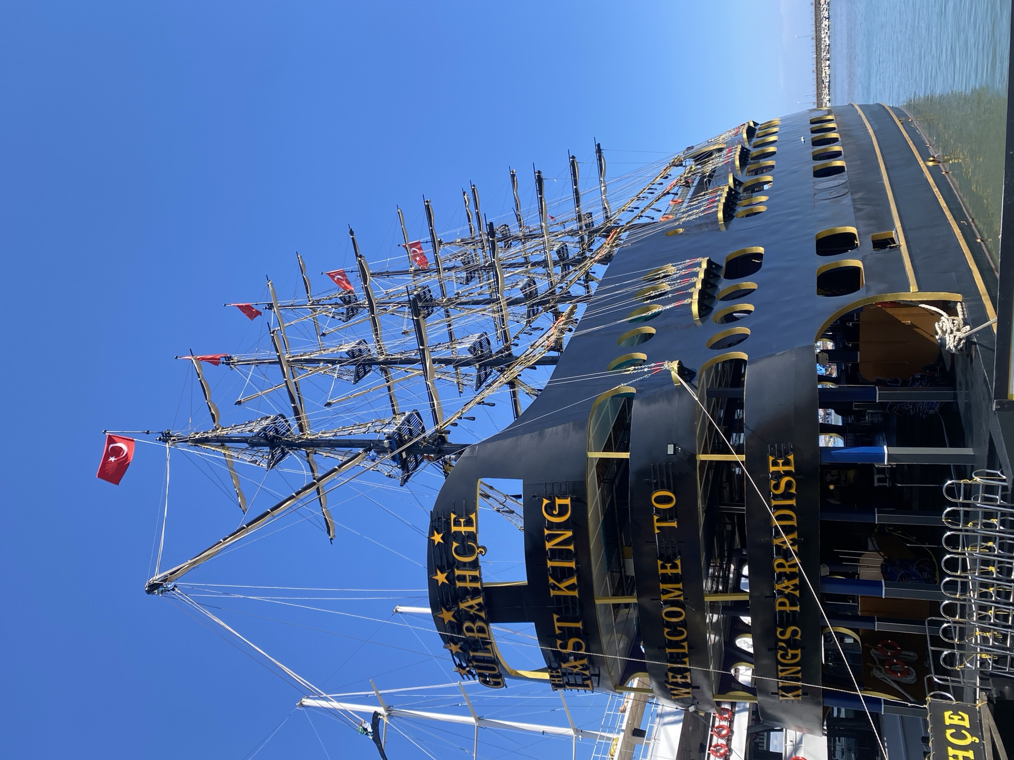 The image shows the "Gülbahçe The Last King" pirate ship in Alanya, Turkey, docked in the harbor. The ship is black with gold trim around its rectangular windows and signage. The signage reads "Gülbahçe The Last King, Welcome to King's Paradise." The ship has multiple masts with Turkish flags flying from the top. In the background, the harbor and the city of Alanya are visible. The sky is clear and blue.