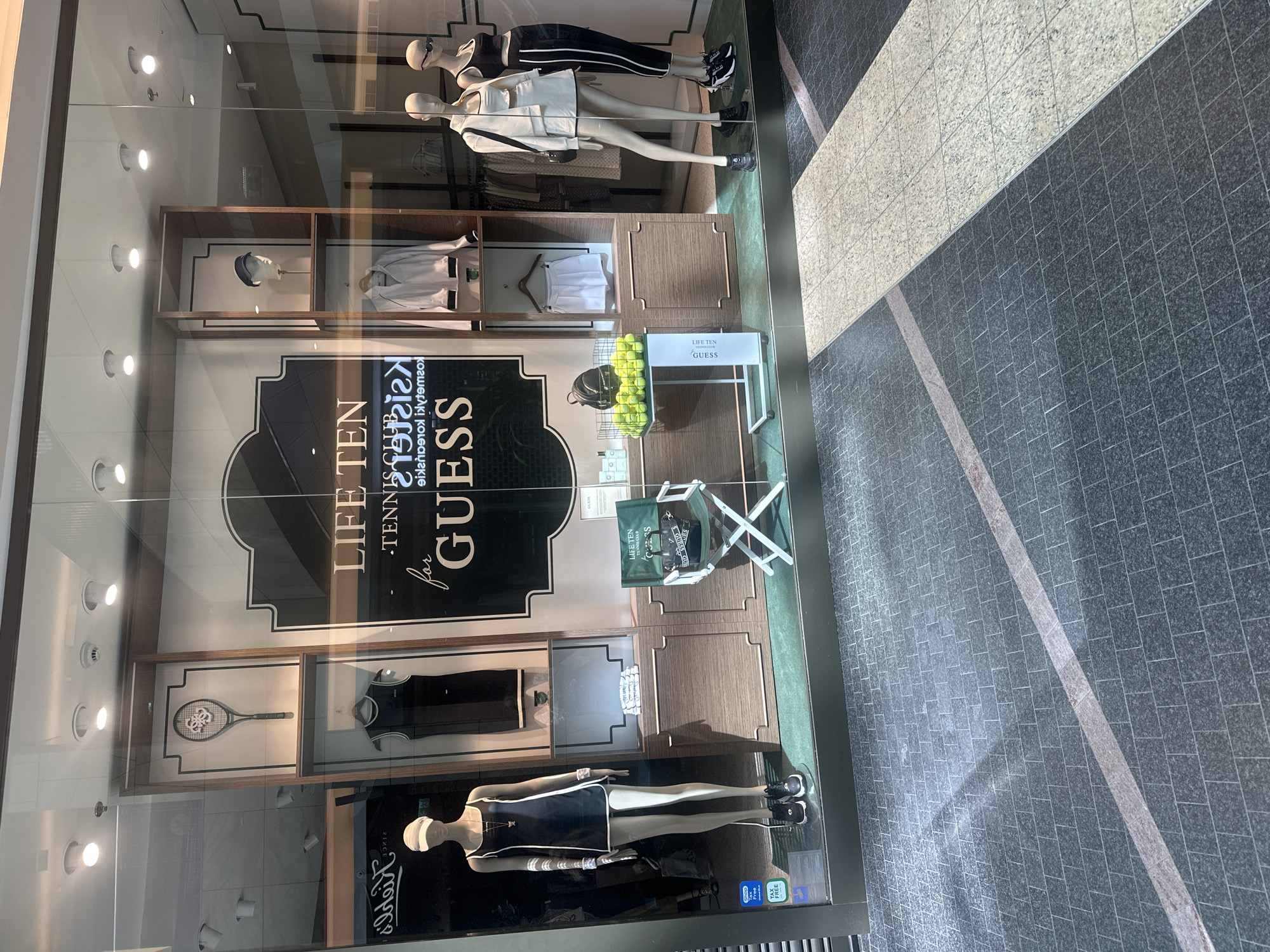 A store window display features mannequins dressed in Guess sportswear, with a tennis theme. One mannequin on the left wears a black and white tennis dress. In the center, a green folding chair with a Guess logo and a display of tennis balls and a backpack are present. To the right, two mannequins are displayed: one in a white tennis outfit and another in a black athletic outfit. Behind the mannequins, clothing racks and shelves with merchandise are visible. The store's branding, "LIFE TEN TENNIS CLUB for GUESS," is prominently displayed. The window is part of a retail establishment located in Warsaw, Poland, as indicated by multilingual signage, including a "TAX FREE" sticker on the left side of the display. The floor in front of the display is paved with dark grey and light grey square tiles, with a diagonal red stripe marking a pathway.