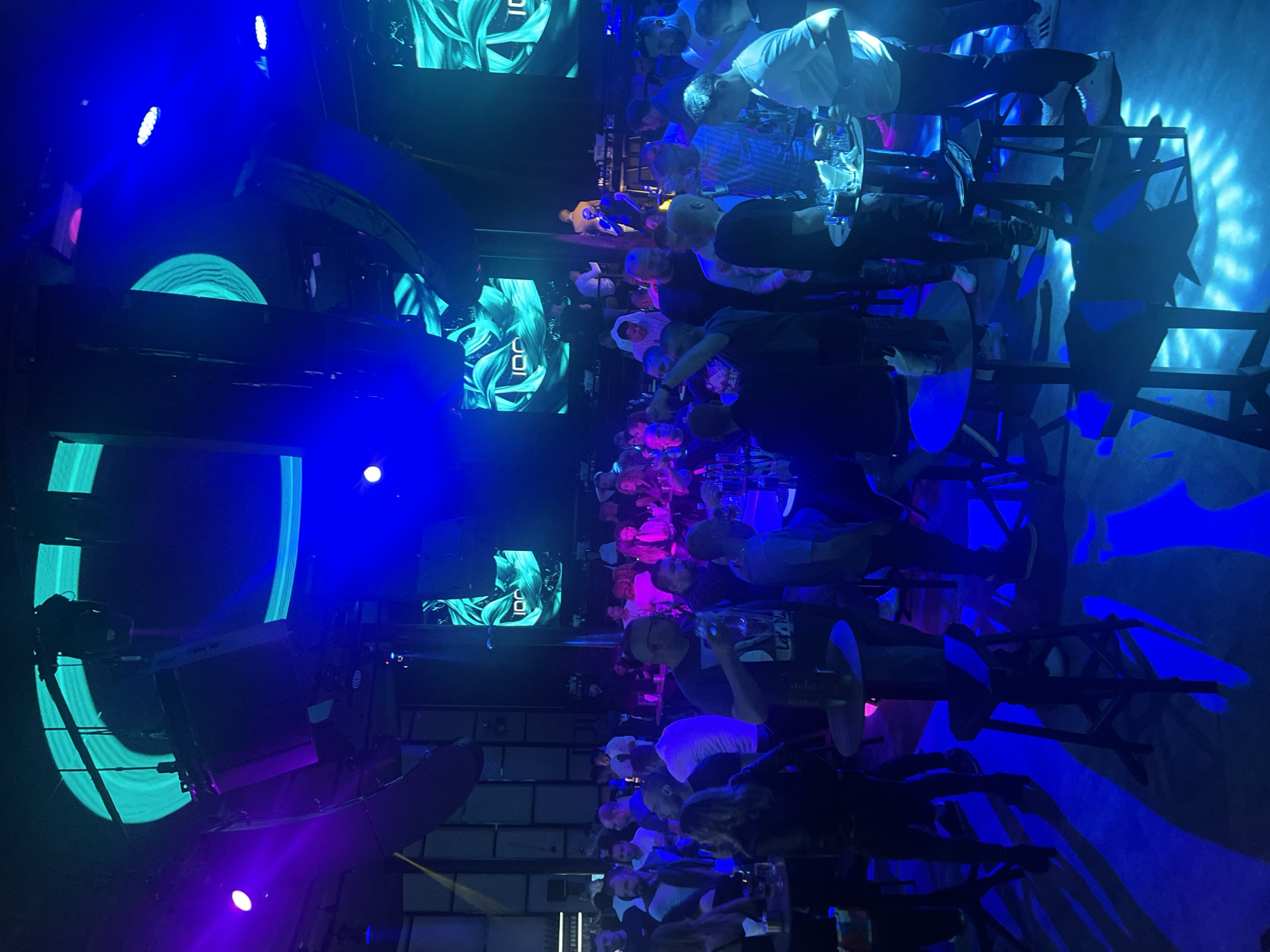 The image depicts an indoor scene, likely a bar or nightclub in Varna, Bulgaria. A large group of people are standing at elevated tables with drinks. The setting is illuminated by blue and purple lighting, with decorative screens displaying abstract designs on the walls and ceiling. Some individuals are interacting with each other, while others appear to be observing the scene. Tall stools surround the tables. The overall atmosphere suggests a lively and social gathering in a nightlife setting.