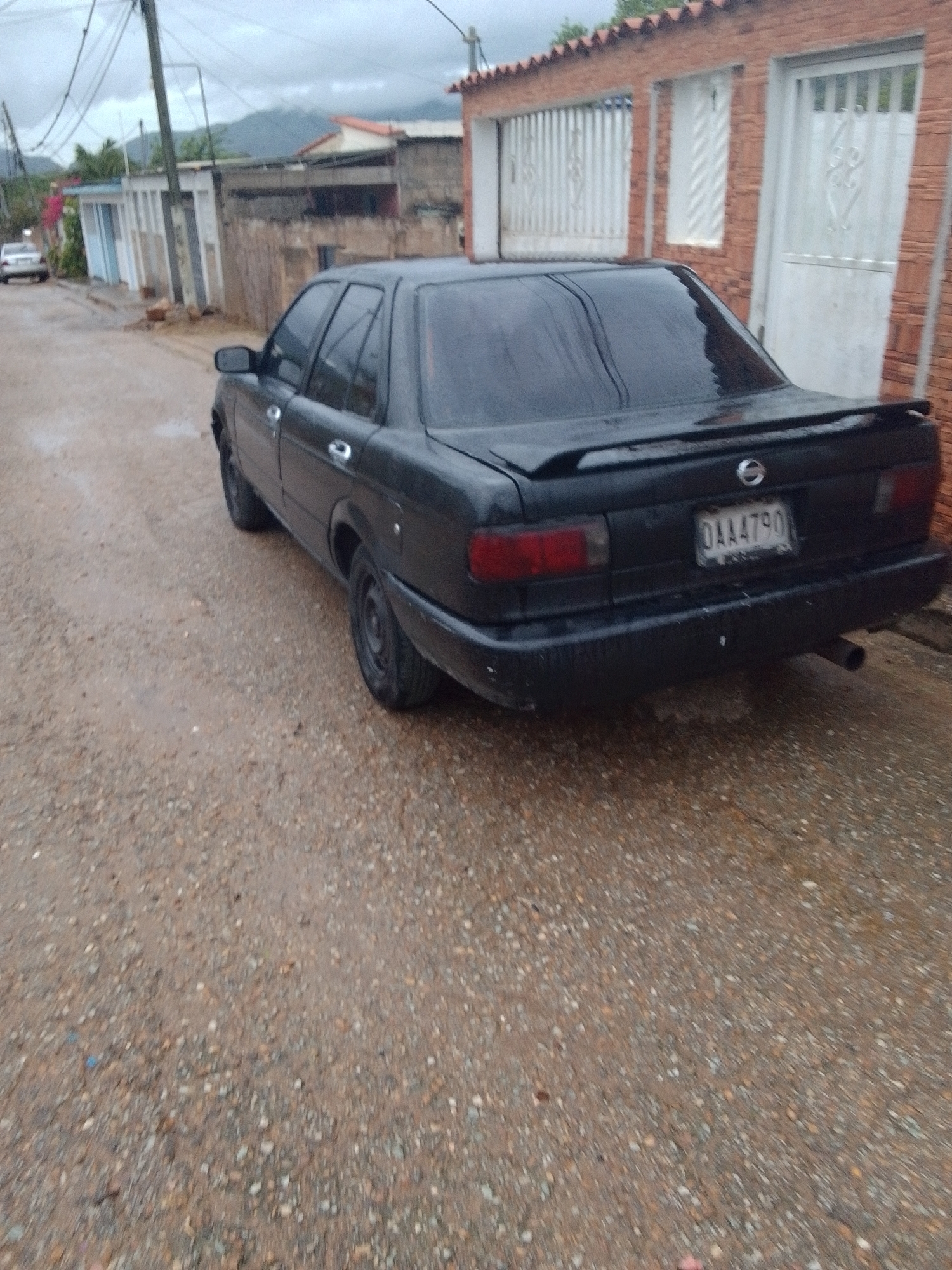 The image shows a black sedan parked on a gravel road in what appears to be a residential area. The car is the primary object in focus, viewed from a slightly elevated rear-quarter angle. It has a spoiler on the trunk and a license plate with the text "OAA4790". The car's windows are dark, and the bodywork appears wet, suggesting recent rain.
The background shows buildings with brick walls and white-trimmed windows and doors. Some of the windows are covered with decorative metal grates. A utility pole with numerous wires stands to the left of the car, indicating the presence of infrastructure. Further in the background, there are more buildings and some greenery, with hills or mountains visible in the distance under a cloudy sky.
The overall scene suggests an overcast or rainy day. The gravel road and the partially wet surfaces contribute to this impression. The environment appears to be a humble, possibly rural or semi-urban setting. There are no people visible in the image. The activity taking place is simply the car being parked, with no indication of any ongoing action. The mood is quiet and perhaps a bit somber due to the weather. The location context of Juan Griego, Venezuela, suggests a tropical or subtropical climate, which aligns with the cloudy weather and potentially humid conditions implied by the wet surfaces.
