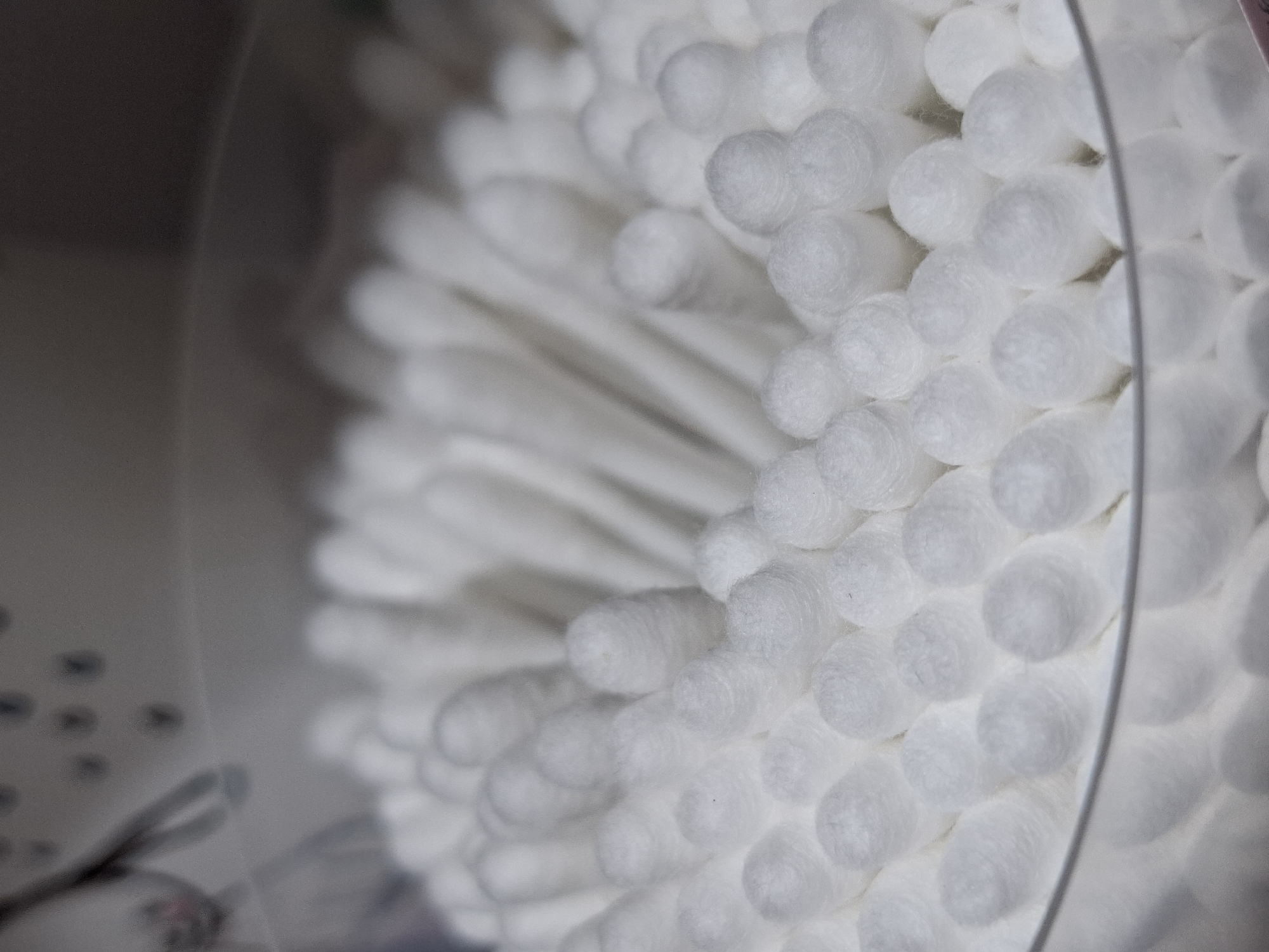 The image provides a close-up view of numerous white cotton swabs, tightly packed within a clear, cylindrical container. The foreground elements, primarily the cotton tips, are in sharp focus, revealing their soft, fibrous texture. The swabs are arranged in a dense, fanned pattern, giving the impression of a full container. A curved, transparent edge, consistent with the rim or side of a plastic container, is visible on the right side of the frame.
The background on the left is heavily blurred and appears dark gray or brown, with indistinct shapes that are too out of focus to identify. No specific activity is depicted; it is a still-life photograph of hygiene products. The lighting is soft and even, highlighting the pure white color of the cotton. There are no identifiable location cues, time of day, or weather conditions visible in the image itself. No text is visible anywhere in the frame.