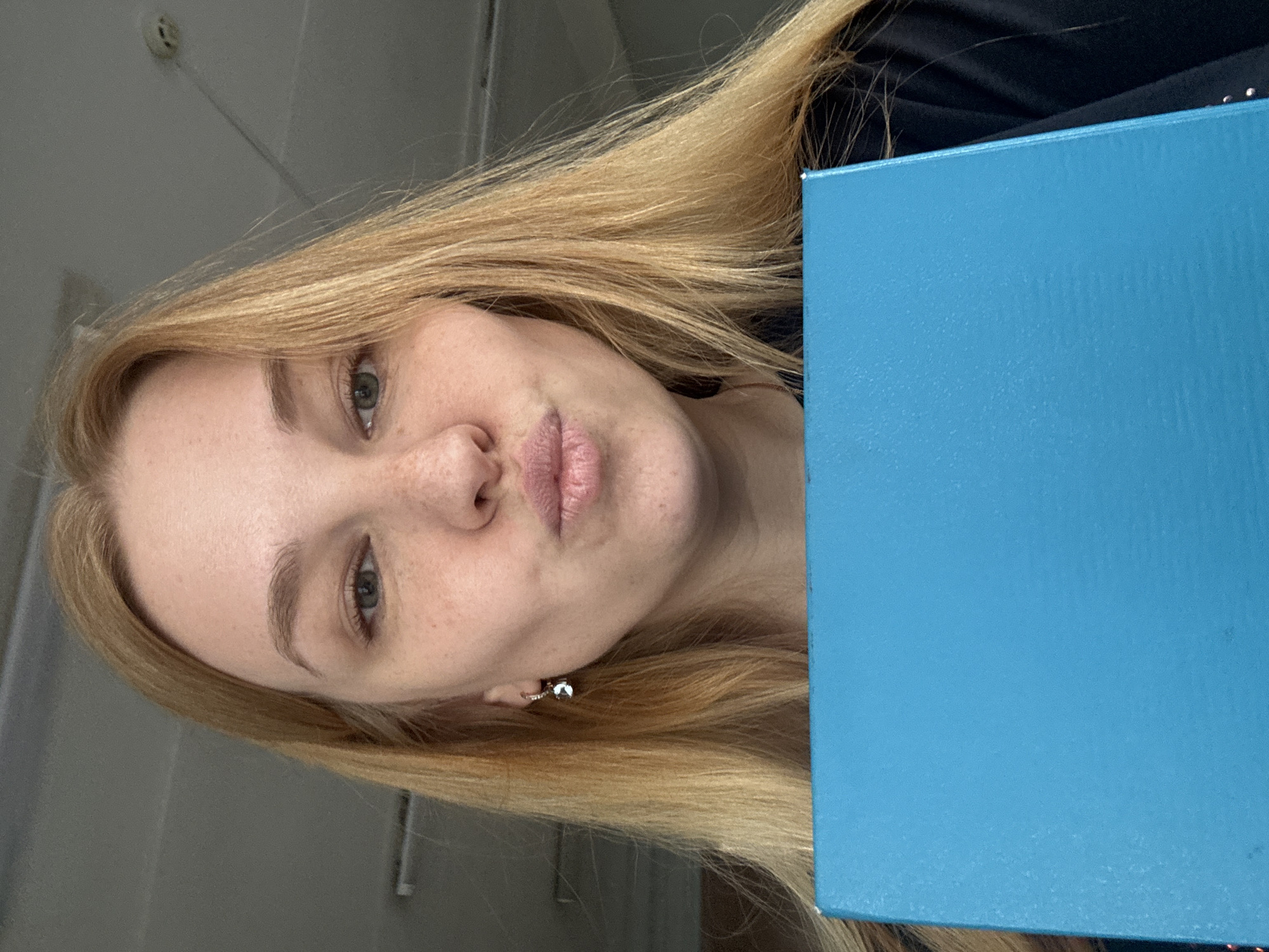 A selfie features a fair-skinned, blonde-haired woman with freckles. She is holding a bright blue, rectangular object, possibly a book or folder, partially obscuring her right side. The woman has light blue eyes and is wearing a small, possibly diamond, earring in her left ear. Her lips are pursed in a kissing expression. The background is a blurry, light grey wall. There is no discernible information that connects the image to Rusanovo, Russia.