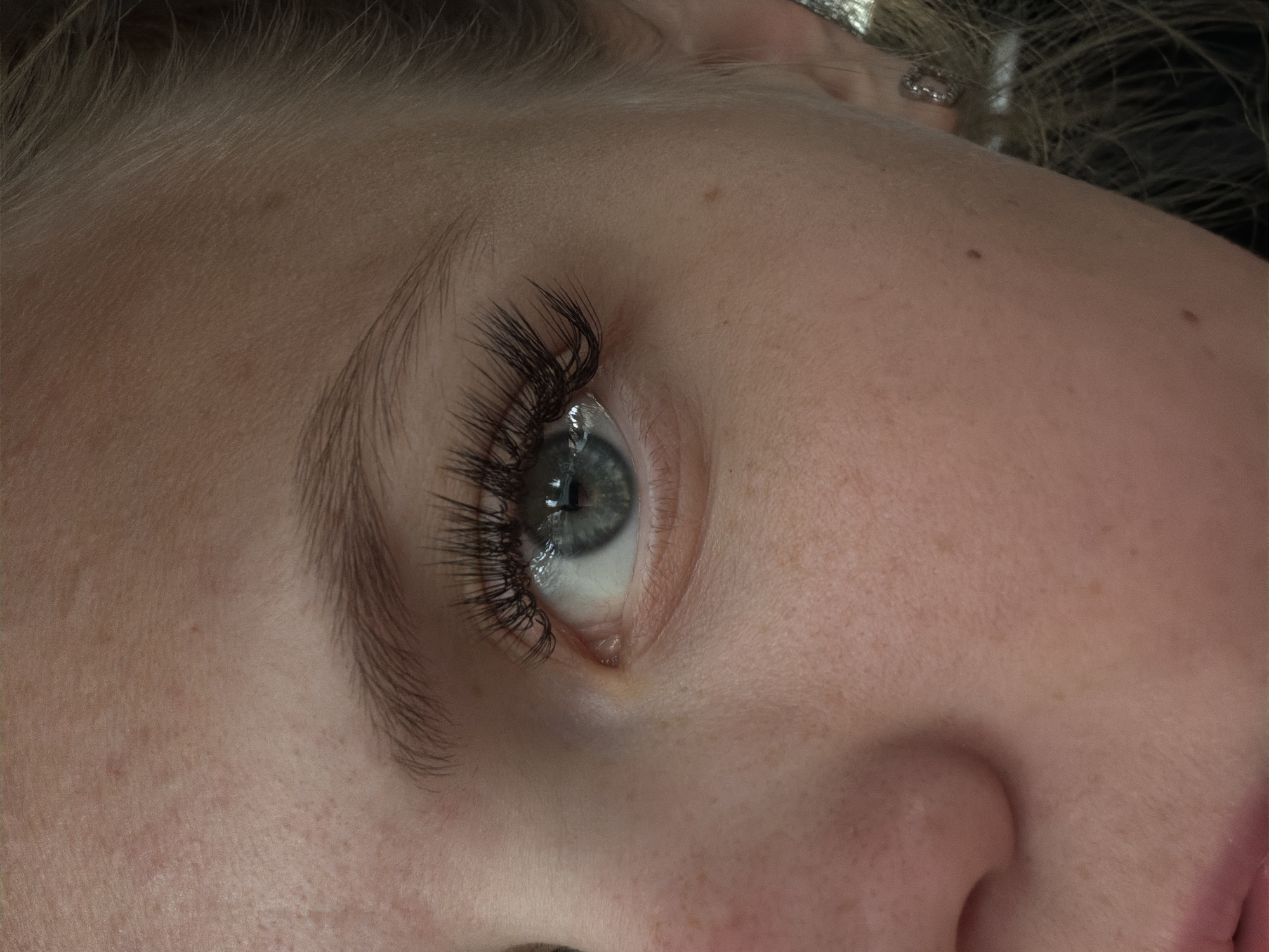 The image is a close-up, high-angle shot of a person's eye and surrounding facial features. The eye is open and looking slightly to the left. The iris is a light blue-green color with visible darker rings and a reflection of what appears to be a light fixture. The person has long, dark, and voluminous eyelashes, likely enhanced with mascara or false lashes. The skin has a fair complexion with visible freckles and some minor blemishes. The eyebrow is dark and well-defined, arching above the eye. Part of the nose, including a nostril and the bridge, is visible on the left side of the frame. The right side of the image shows a portion of the ear with a small, sparkly earring and some blonde hair. The background is dark and out of focus, suggesting the photo was taken indoors with controlled lighting. The image does not provide any information about the city of Chaikovsky, Russia, as the scene depicted is an isolated facial close-up.