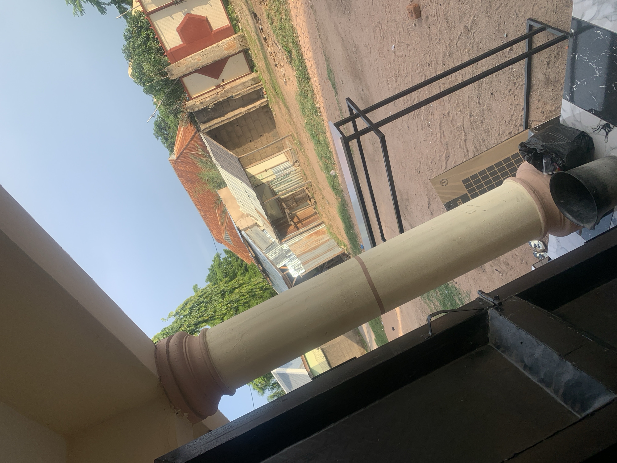 The image captures an outdoor scene in Jauro Yinu, Nigeria, showing a view from under a covered porch. A large, cream-colored pillar with decorative brown bands dominates the left side of the frame, leaning towards the right. To the left of the pillar is a dark-colored doorway or entrance.
Beyond the porch area, the environment transitions to a sandy, uneven ground with patches of green grass. In the mid-ground, several structures are visible. One appears to be a small building with a corrugated metal roof, constructed with brick or concrete blocks. Adjacent to it is another structure, possibly also a dwelling or shop, with walls made of rough materials and a metal roof. Further in the background, a taller building with a distinct red-tiled roof is partially visible. A decorative wall, painted cream with bold red geometric patterns, stands to the right of these structures.
Several objects are scattered on the ground in the foreground and mid-ground. These include a black trash bag, a dark-colored bucket, a flat, rectangular object with a grid pattern (possibly a solar panel), and a metal shelving unit with black frame. The overall impression is one of a somewhat informal and developing urban or semi-urban setting. The lighting suggests it is daytime, likely late afternoon due to the angle of the sun casting soft shadows. The sky is clear and blue, with some trees visible in the distance.