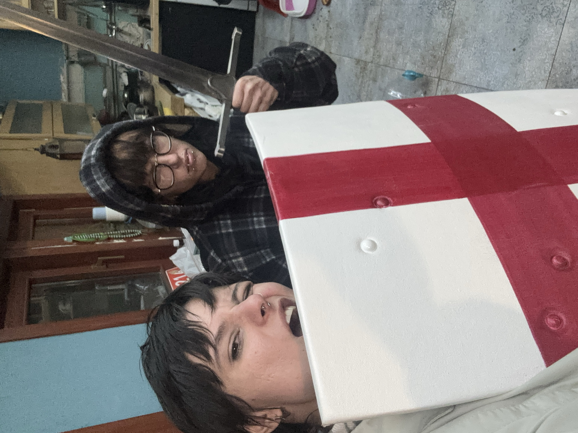 Two individuals are visible in the image. One is wearing glasses and a plaid hoodie, holding a sword. The other individual has black hair and facial piercings. A white and red cross is prominently displayed in the foreground. The location is a cluttered indoor space, possibly a residential area. The image does not provide enough detail to determine if it is in Verinsko, Bulgaria.