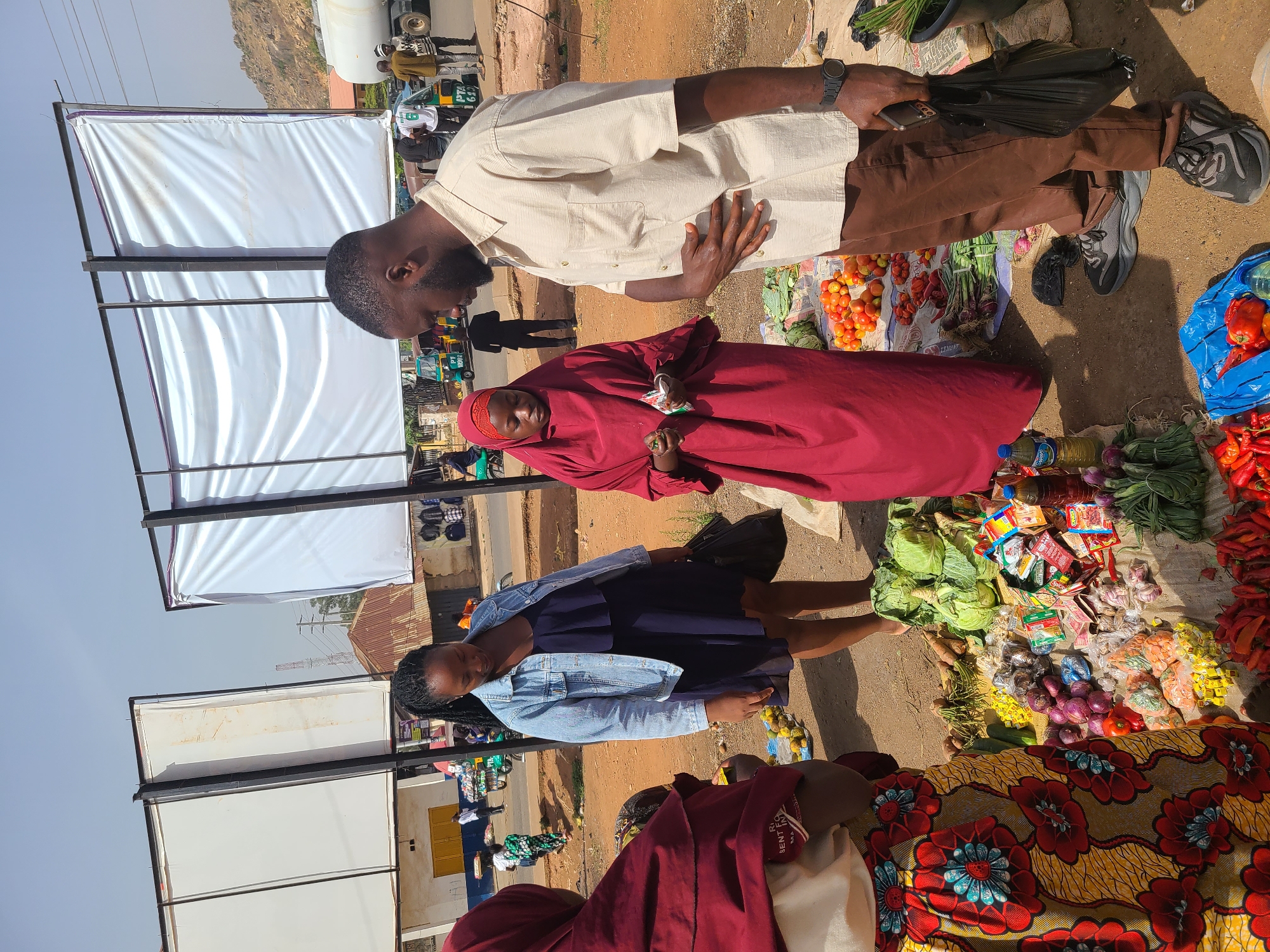 The image depicts an outdoor market scene in Jos, Nigeria. Three individuals are visible: a man in a light beige shirt and brown pants, a woman in a red headscarf and dress, and a woman in a blue denim jacket and dress. The man appears to be inspecting produce, while the woman in red is holding what appears to be a small package of goods. The other woman is standing near a display of vegetables. Various vegetables, including tomatoes, onions, peppers, and leafy greens, are spread on the ground for sale, arranged on a colorful patterned cloth. In the background, there are white fabric canopies on a metal frame, and a street scene with cars and buildings. The image suggests a typical day at a local market in Jos.