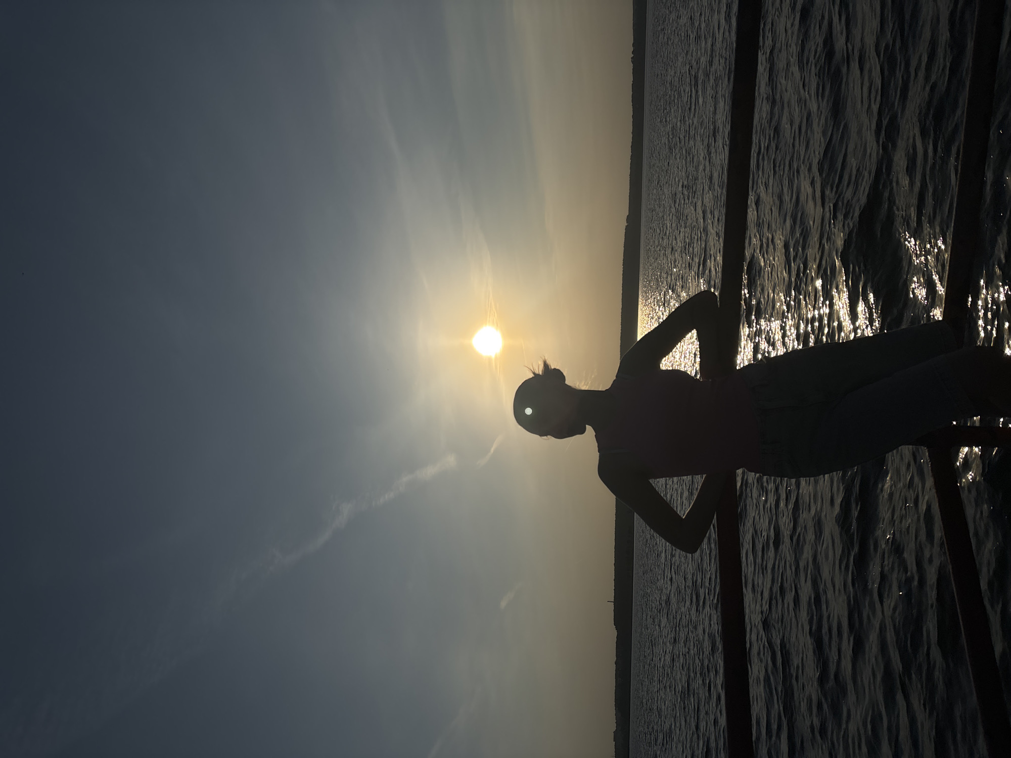 The image depicts a silhouette of a person standing on a structure over water at sunset. The sun is positioned directly behind the person's head, creating a halo effect. The water reflects the sunlight, creating a shimmering effect. Vertical poles are visible, possibly part of a bridge or pier. The city of Perm, Russia is not identifiable from the details in the image.