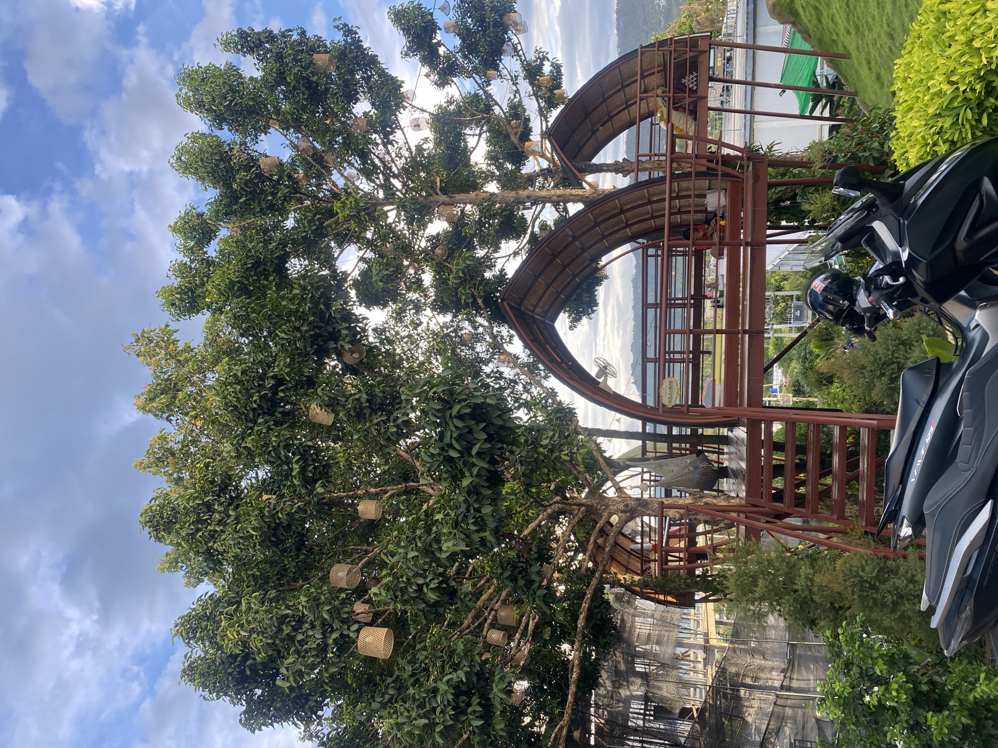The image depicts a scene in บ้านบางรักษ์, Thailand, focusing on a large tree adorned with woven baskets, positioned next to a two-story brown metal structure with curved roof-like elements. A black and silver motorcycle is partially visible on the right side of the frame, obscuring a view of the surrounding area. The background includes hints of a waterfront or bay, visible through the metal structure, with distant landforms. The sky is blue with scattered clouds. The ground is vegetated with greenery.