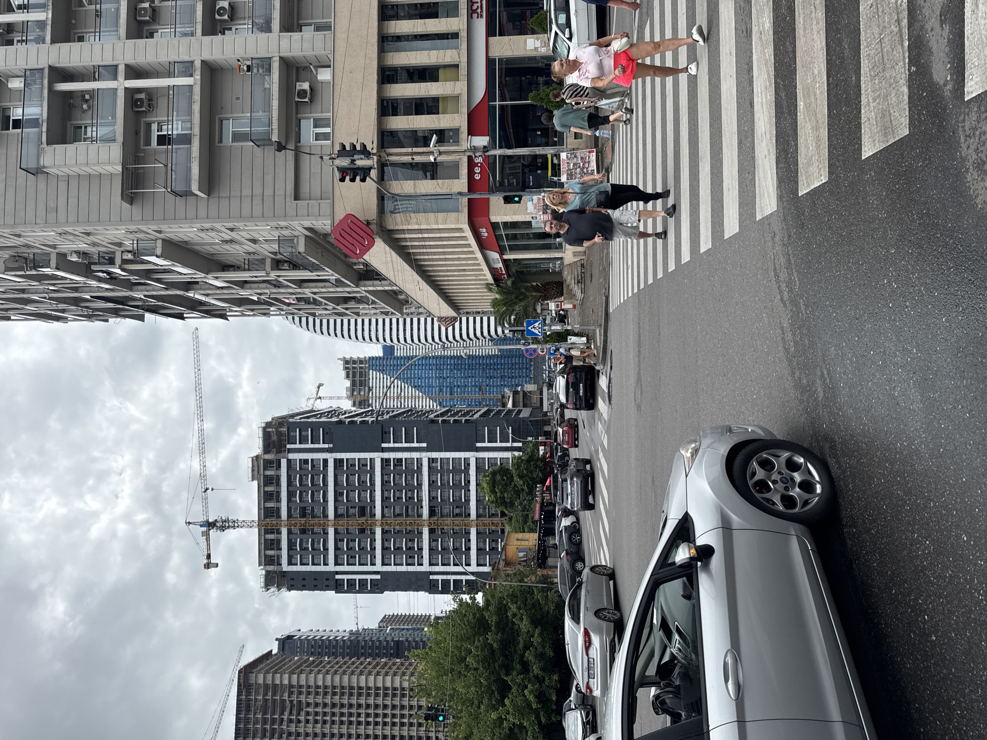 The image depicts a street scene in Batumi, Georgia, likely near the Batumi Boulevard area. In the foreground, a silver Ford Fiesta is partially visible, facing left. Across the street, a pedestrian crossing is occupied by several individuals. To the right of the crossing, a woman in a pink top and shorts walks away from the camera, and a man and woman walk towards the camera. Behind them, other pedestrians are visible.
The street is wet, suggesting recent rain. Traffic lights are present, with one showing a green light for vehicles. Several cars are on the road, including a silver sedan on the left and a black SUV turning.
The background is dominated by modern high-rise buildings under construction, evident by cranes and scaffolding. Some completed buildings also line the street, featuring balconies and air conditioning units. One building displays a large red cube logo. The sky is overcast with grey clouds. The overall impression is of a bustling urban environment undergoing development.
