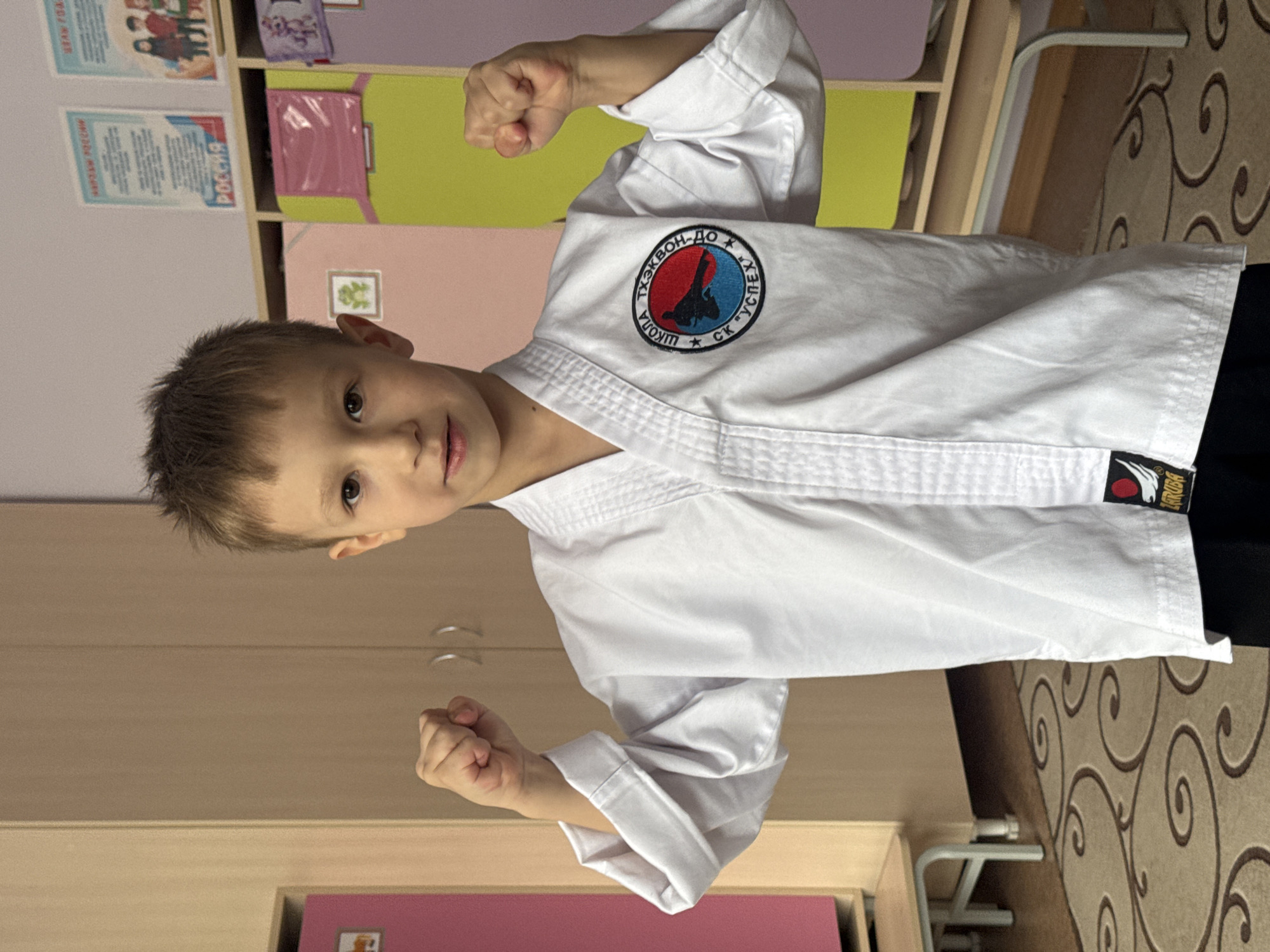 A young boy, approximately 6-8 years old, is depicted indoors, looking directly at the camera with a determined and confident expression. He has short, light brown hair and dark eyes. He is dressed in a crisp white martial arts uniform (gi). On his left chest, there is a prominent circular patch featuring a red and blue design with a stylized figure in a martial arts pose. The patch is inscribed with Russian text: "ТХЭКВОН-ДО" (Taekwon-Do) at the top, "ШКОЛА" (School) on the left, and "СК "УСПЕХ"" (SC "Uspekh," meaning Sports Club "Success") on the right.
The boy strikes a pose, with both arms bent at the elbows and fists clenched and raised, showcasing readiness or strength. A small black tag with the white brand name "ZARUBA" and a red sun logo is visible on the lower part of his right sleeve.
The background shows light-colored wooden or laminate shelving units, possibly part of a cabinet or storage system. Several papers or posters with Russian text are affixed to the back of these shelves; one visible section reads "РОССИИ" (Russia). The floor is covered with a patterned carpet or rug featuring swirling brown and beige designs. A light-colored metal object, possibly a part of furniture, is visible in the lower right. The setting suggests an indoor training facility, a dojo, or a room within a school or youth center in Chaikovsky, Russia.