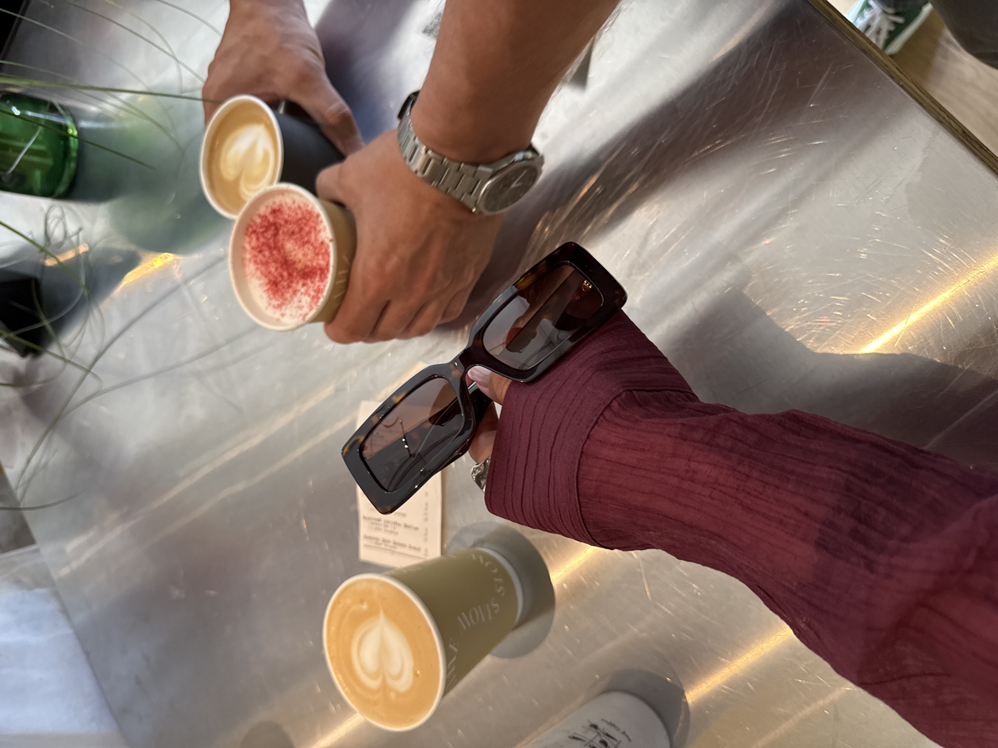 In Esto-Sadok, Russia, a close-up shot captures a stainless steel table surface with three coffees, a pair of tortoiseshell sunglasses, and a wristwatch. The foreground features a hand wearing a deep red textured long-sleeved shirt, holding the sunglasses. To the left, a light green paper cup displays latte art. In the center, a beige cup is topped with red sprinkles. To the right, a dark gray cup holds latte art and is being held by another person's hands. A silver wristwatch with a black face is visible on the wrist of the person holding the dark gray cup. The table surface exhibits numerous scratches and reflections.