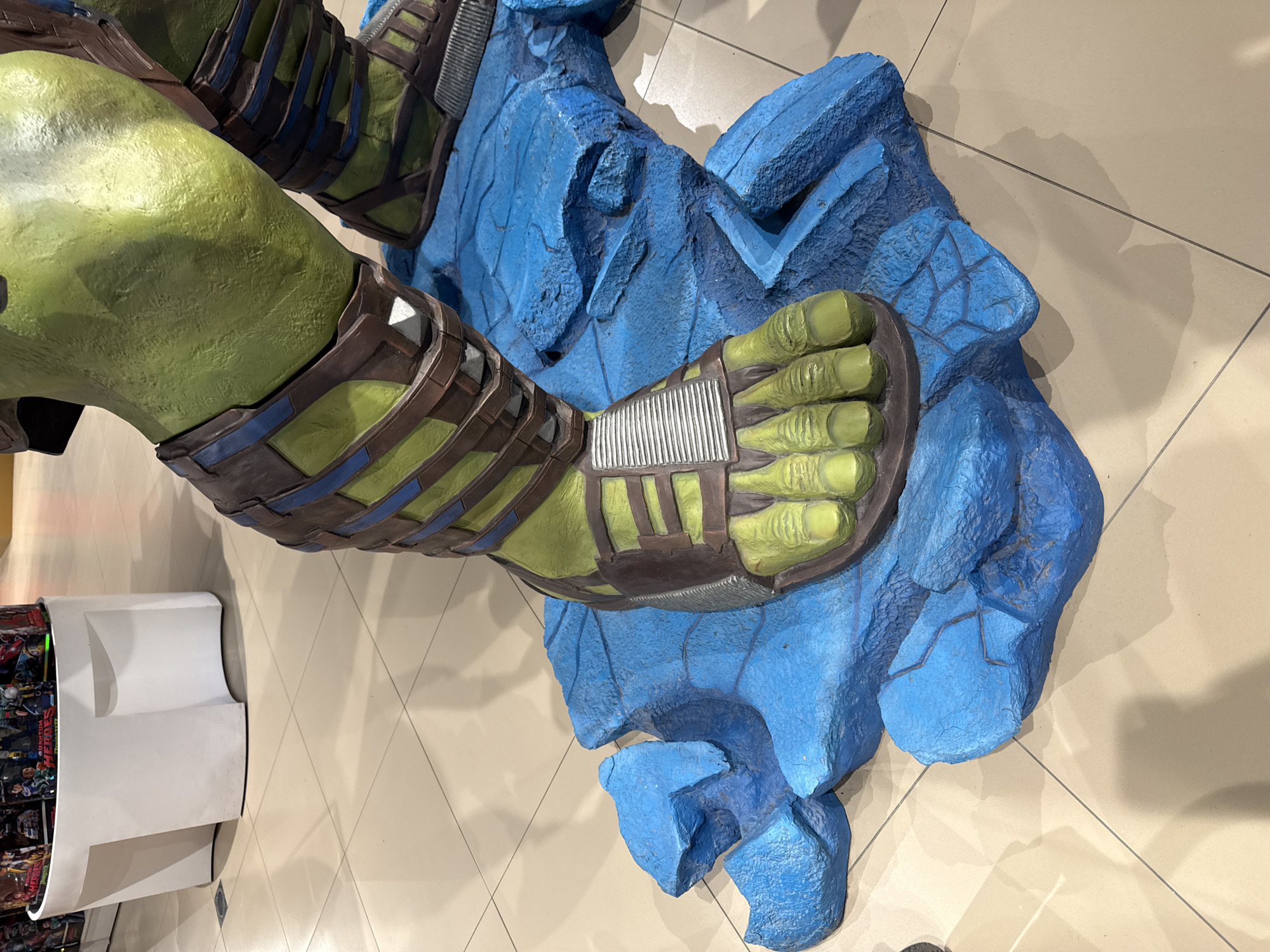 The image shows a partial view of a Hulk figure, specifically its lower leg and foot, standing on a sculpted blue rock formation. The Hulk's leg is green, adorned with brown and blue armor-like straps. His toes are also green and appear to be wearing sandals. The blue rock base has a textured surface. The figure is situated on a light-colored tiled floor. A white display stand with various printed materials is visible to the left of the figure. The location is likely indoors, possibly a retail store or exhibition related to comic books or superhero-themed merchandise. While a specific location in Moscow, Russia cannot be definitively determined from this image alone, this kind of displays are common in shopping malls or theme-related stores in larger cities like Moscow.
