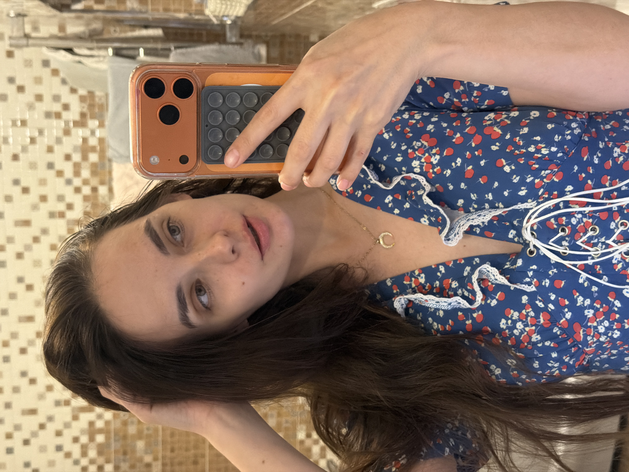 A young woman is taking a selfie in a bathroom. She is wearing a blue floral dress with white lace trim and red flower accents. The dress has a lace-up detail on the front. She is holding an orange phone case with a pop socket attached. Her dark hair is long and wavy, and she has light-colored eyes and fair skin. The background appears to be a tiled wall and a mirror. The lighting suggests it might be daytime, though the exact time is unclear. The woman's expression is neutral.