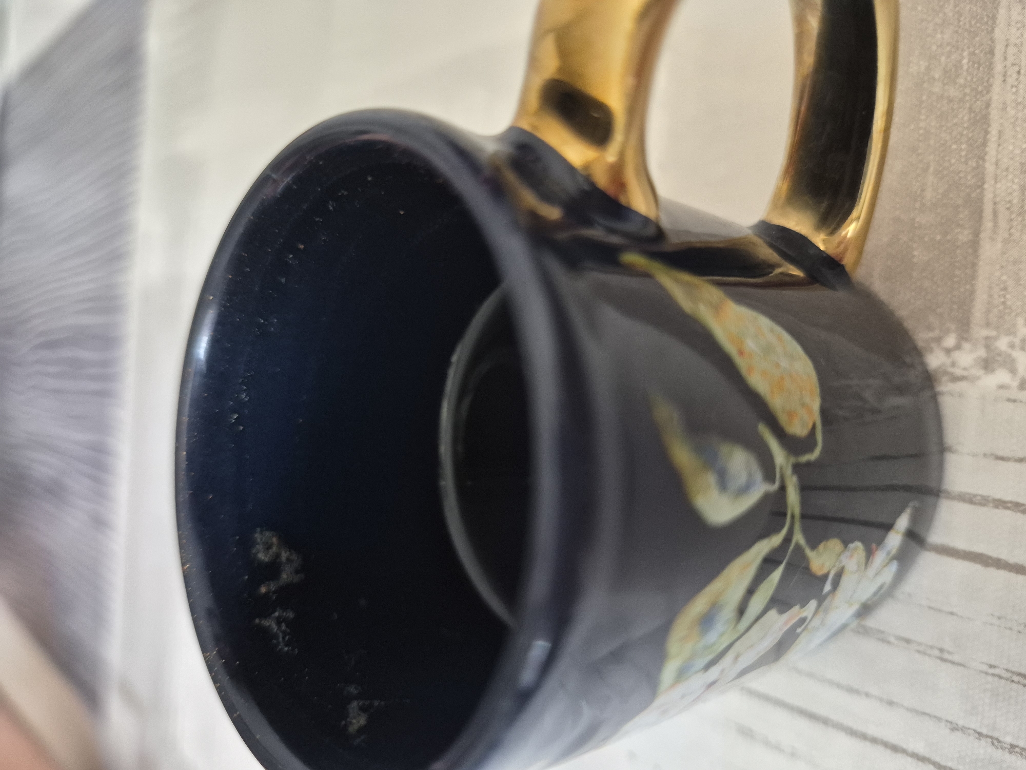 A close-up, eye-level shot shows the inside and side of a dark blue mug with a gold handle, decorated with light-colored floral motifs. The mug is placed on a surface with a striped pattern. The background is blurred, showing a light-colored surface. The lighting is natural and illuminates the mug from the front. The scene is set in an indoor environment.