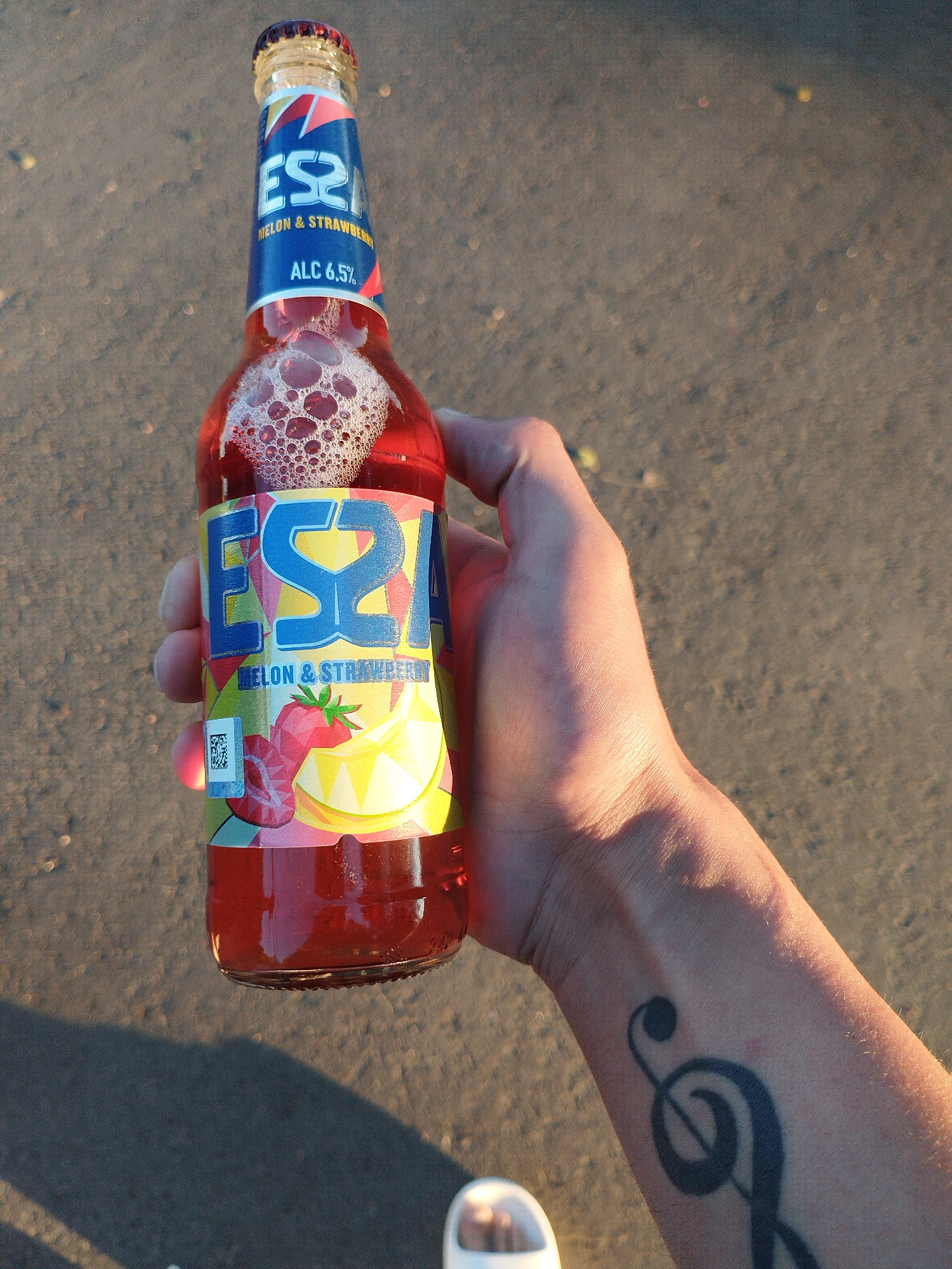 A person's hand holds a bottle of pink liquid with a label that reads "ESA" and "MELON & STRAWBERRY" with "ALC 6.5%" also visible. The bottle has foam at the top. The background is an asphalt-like surface. A tattoo of a treble clef is visible on the person's forearm. The lighting suggests it is either sunrise or sunset.