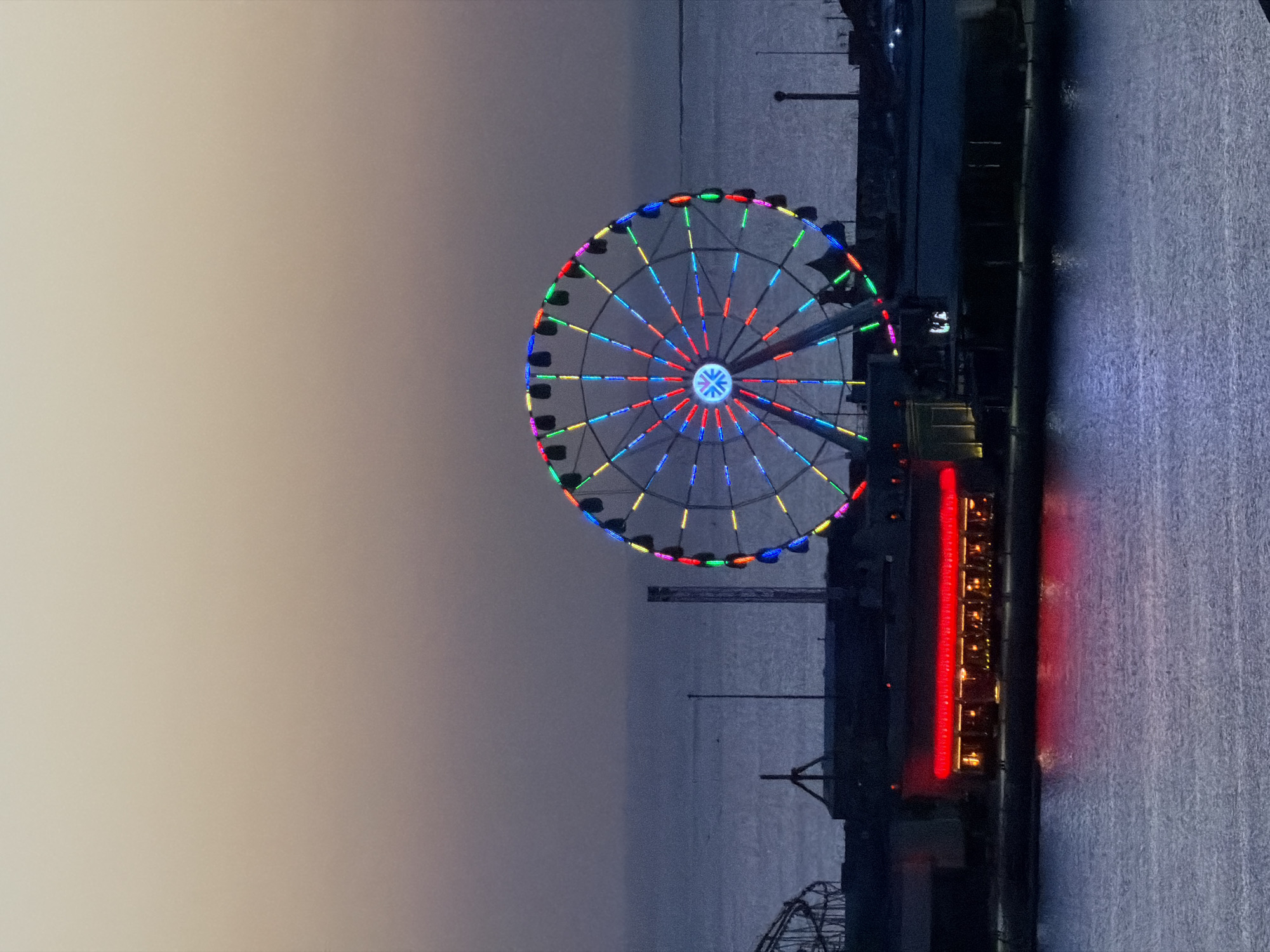 An outdoor, vertical, medium shot captures a Ferris wheel at dusk against a twilight sky, positioned over a body of water. The Ferris wheel, illuminated by a rainbow of multi-colored lights, is the primary subject. Red neon lights outline a building with visible windows and interior lighting, situated below the Ferris wheel. The water in the foreground reflects the lights from the building. The sky transitions from a pale yellow-beige at the top to a darker grey-purple at the bottom. In the background, a silhouette of a roller coaster is visible to the left of the Ferris wheel, along with other structures and what appears to be distant land or a coastline. The scene is set in an unspecified urban waterfront location at dusk.