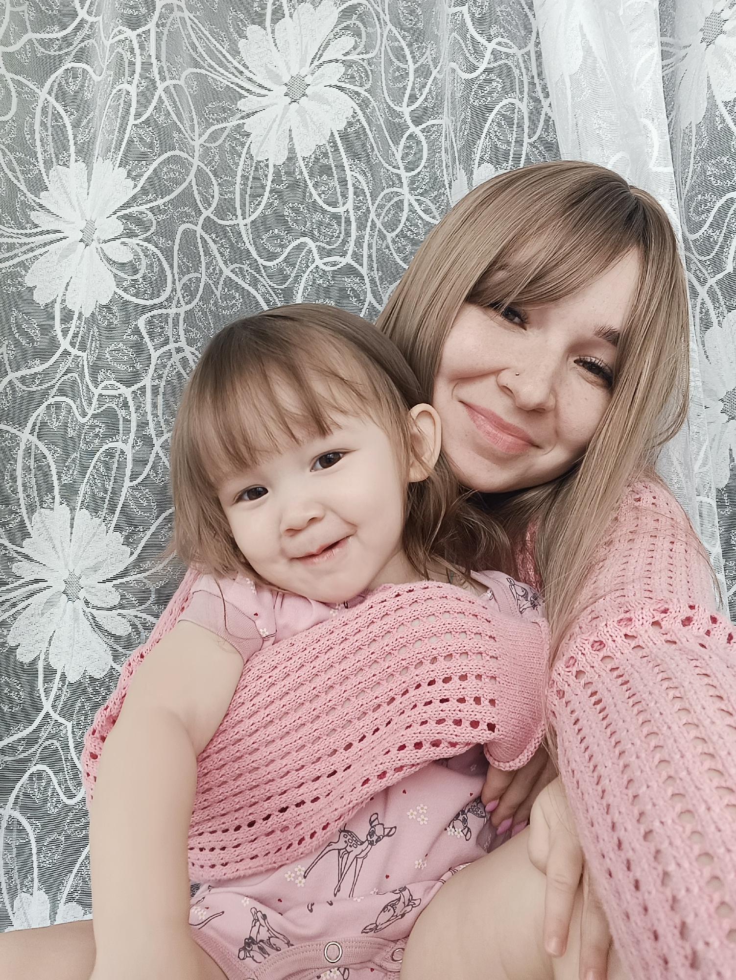 The scene depicts two individuals, an adult female and a young child, indoors in what appears to be a domestic setting. The adult female, with light brown hair and bangs, a small nose piercing on her left nostril, and freckles, wears a light pink, open-knit sweater. Her fingernails are painted pink. She is positioned behind and to the right of the child, smiling at the camera. The child, a toddler with brown hair and bangs, wears a light pink romper or bodysuit featuring a pattern of deer and flowers. A light pink, open-knit garment is draped over the child's shoulders. The child is also smiling at the camera. The interaction suggests a close relationship, with the adult embracing or holding the child. The background consists of a white, sheer curtain with a repeating floral and swirling pattern, positioned in front of a darker surface. No specific landmarks or unique identifiers are visible to pinpoint a precise location within the city of Izhevsk, Russia.