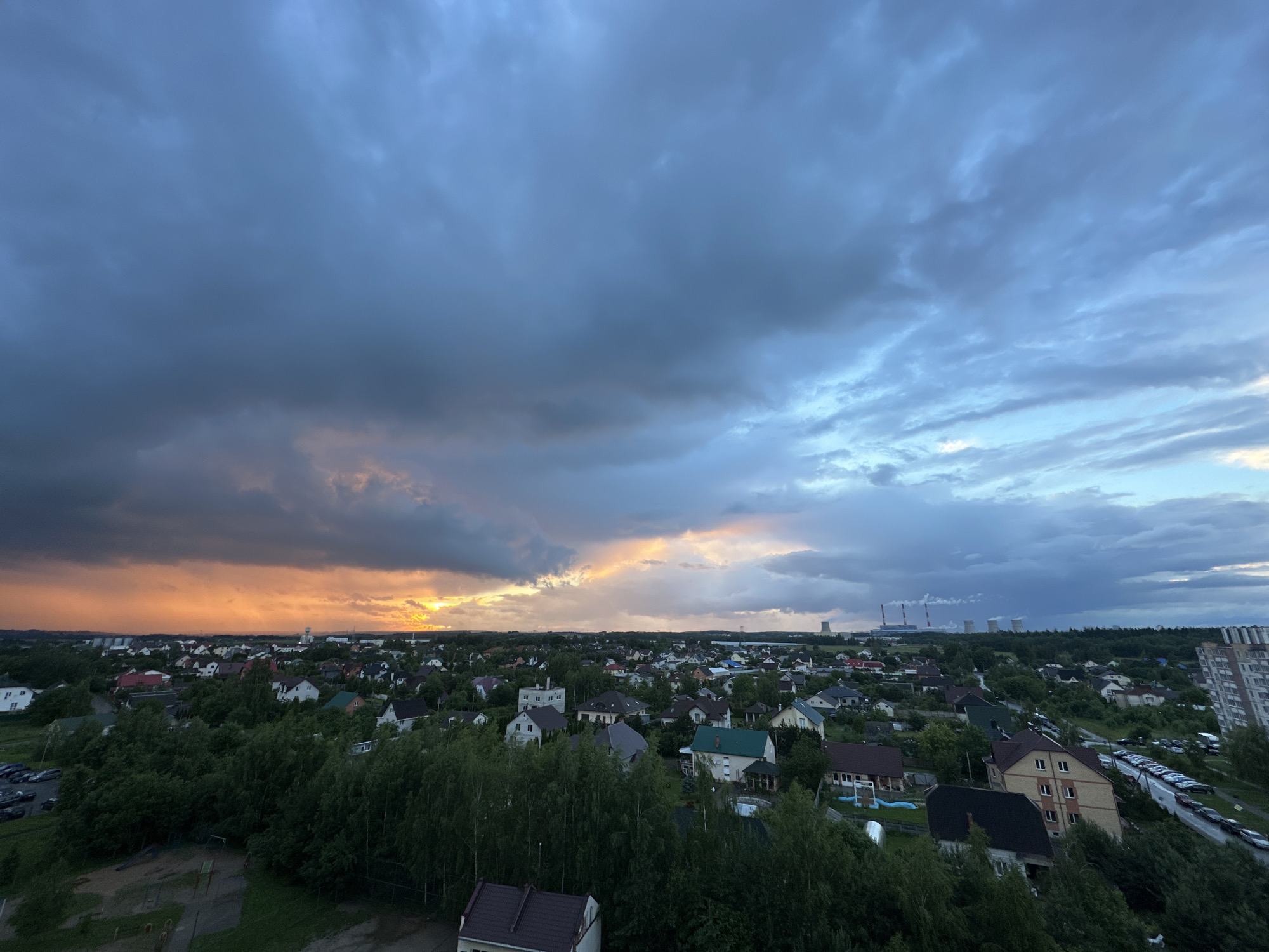 An aerial, wide-angle view depicts a suburban landscape in the vicinity of Shchomyslitsky Sel'skiy Sovet, Belarus, under a dramatic sky. The horizon is dominated by a vast expanse of clouds, transitioning from a dark, ominous gray on the left to lighter blues and whites on the right. A vibrant band of orange and yellow light breaks through the clouds on the left, suggesting the setting or rising sun. Below the sky, a densely packed residential area with numerous houses, varying in architectural style and color, is visible, interspersed with abundant green trees. A network of roads with parked cars is present, particularly along the right side of the image. In the distance, industrial structures with smokestacks are faintly discernible against the horizon. The foreground shows a section of a playground and more houses with dark roofs, indicating a residential neighborhood. The overall impression is one of a transition in weather, with the potential for rain indicated by the dark clouds.