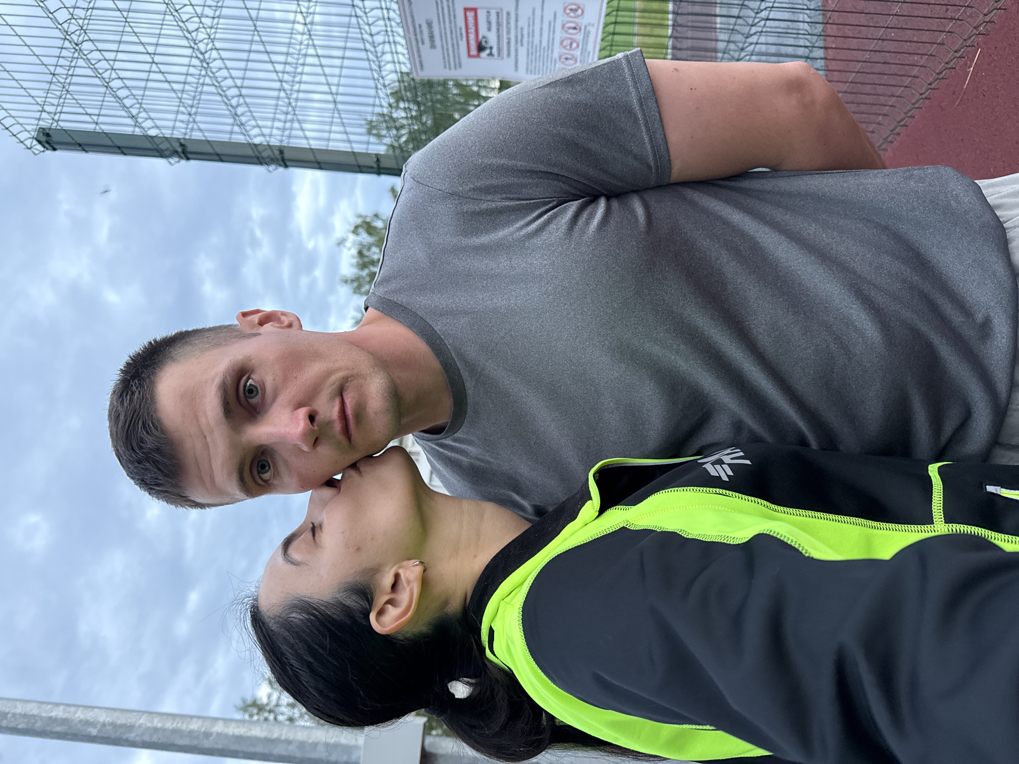 A man and a woman are kissing in what appears to be an outdoor sports facility in Chaikovsky, Russia.
The man is fair-skinned with short, dark hair and is wearing a gray t-shirt. His eyes are looking directly at the camera.
The woman has dark hair pulled back in a ponytail and is wearing a black jacket with neon green accents. She is kissing the man's cheek.
They are standing near a chain-link fence. Behind them, there's a sign with warning symbols and text in Russian. The ground appears to be a red, rubberized sports surface. The sky is cloudy.