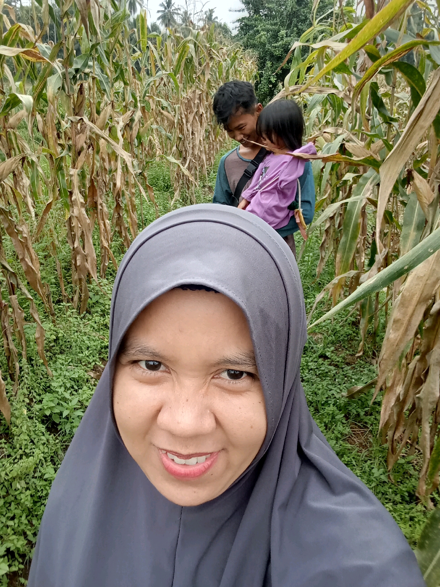 In a cornfield in Campago, Indonesia, a woman in a grey hijab is pictured in the foreground, smiling directly at the camera. Behind her, slightly out of focus, a young man carries a small child wearing a purple shirt. The corn stalks are tall and dry, with some green foliage visible at the base of the plants. The sky is overcast.