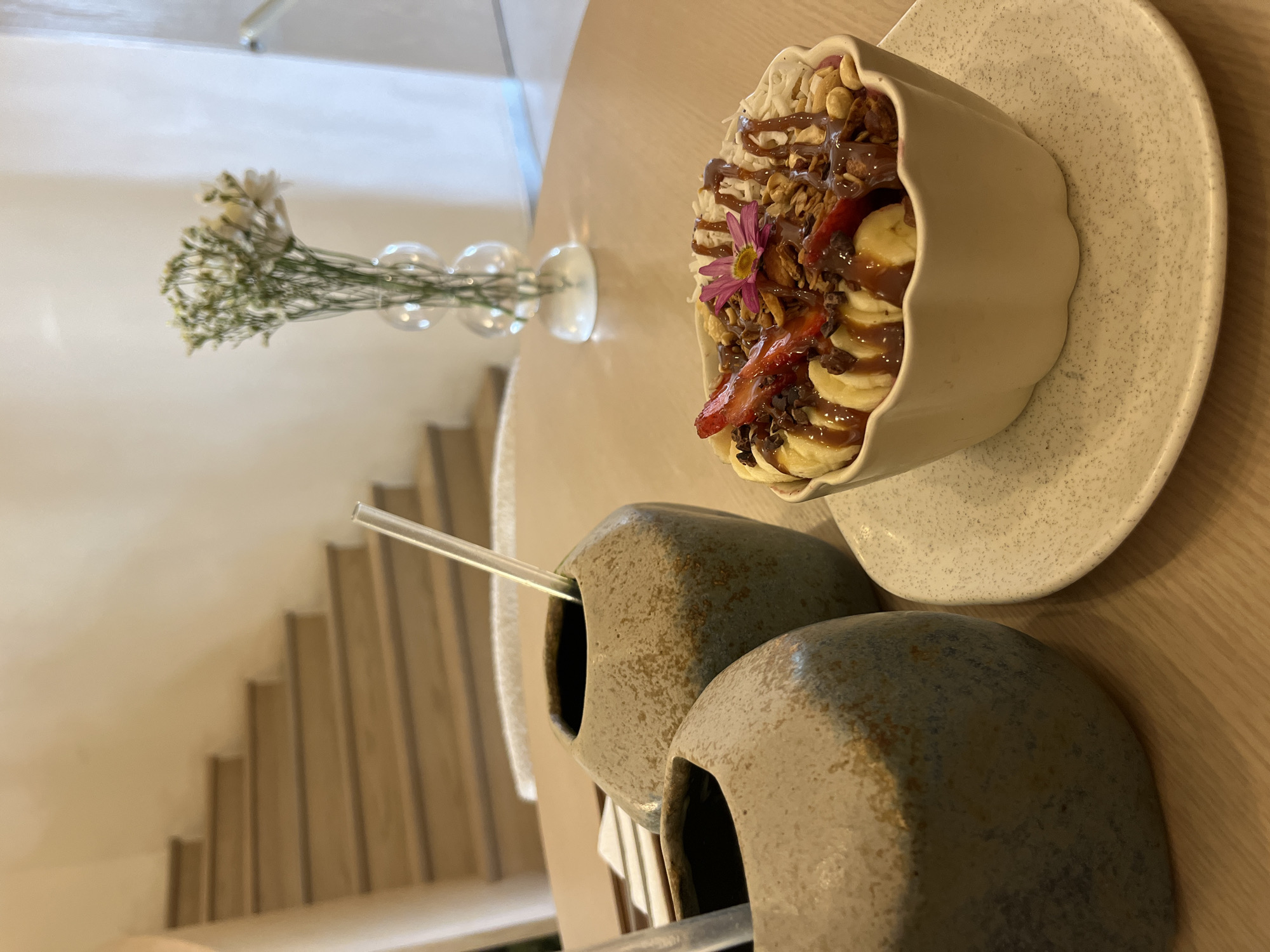 The image shows a close-up of a table setting, likely in Puebla, Mexico, featuring a decorative bowl filled with a dessert. The dessert appears to be a smoothie bowl, containing sliced bananas, strawberries, granola, possibly shaved coconut, and drizzled with chocolate or caramel sauce. A purple flower is placed on top as a garnish. The bowl rests on a speckled, off-white plate.
In the foreground, there are two ceramic vessels with straws, possibly holding drinks. Behind the table setting, a vase with white flowers is visible. In the background, there is a glimpse of a staircase in a light-colored interior. The setting suggests a café or restaurant environment.