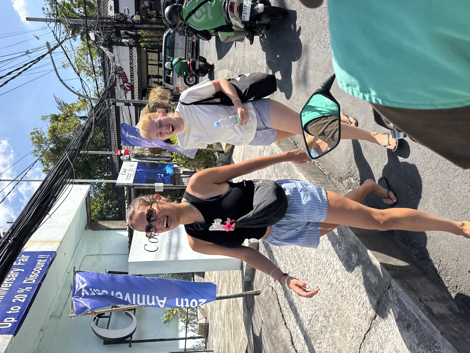 The photograph captures two smiling women standing on a street in Kuta, Indonesia. One woman has blonde hair and wears a white shirt and striped shorts, holding a water bottle. The other woman has dark hair and sunglasses and is wearing a black tank top and striped shorts with a pink hair clip. A motorcycle mirror is visible, with the person holding the camera on a motorcycle.
Behind the women, there are scooters with the "gojek" logo, a parked white van with the word "BURGERS" on it, and a building with signs and banners advertising an "Anniversary Fair" with discounts. Overhead, a dense network of electrical wires stretches across the sky. The location appears to be a bustling commercial area in Kuta, Indonesia.