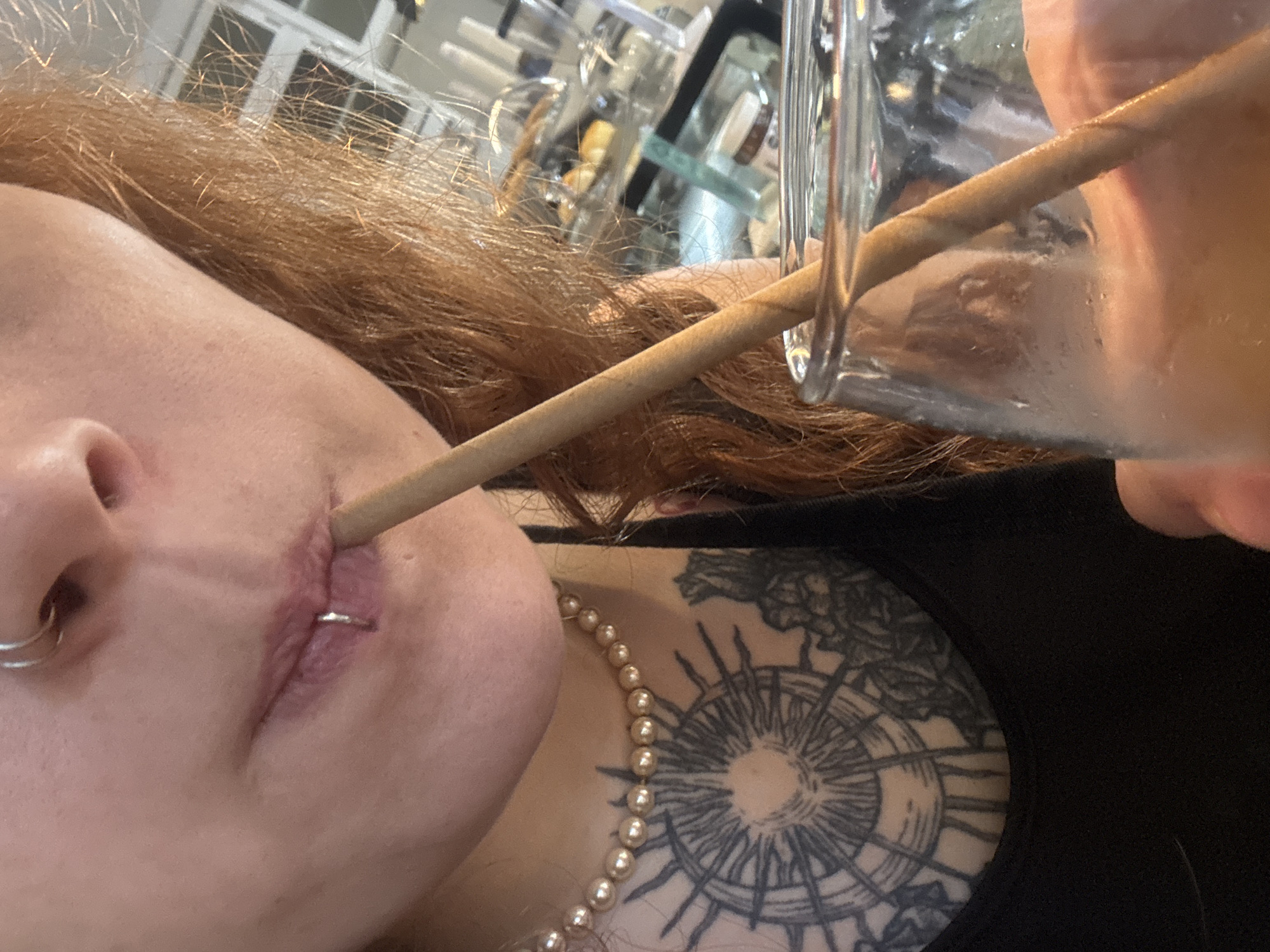 A close-up, tilted view shows a person drinking from a glass with a straw, set in an indoor environment in Buenos Aires, Argentina. The person's profile is visible from the nose down to the upper chest. They have red or auburn hair, part of which is visible along the top of the frame and in the background. A silver nose ring is visible on the left side of their nose. A thin, light-brown straw, possibly made of wood or paper, extends from a clear glass towards their mouth. A silver piercing is visible on their upper lip, and the skin around the piercing and where the straw enters the mouth appears somewhat red or puckered. Around their neck, a string of small, round, golden-colored pearls is worn. On their chest and shoulder, a large, intricate tattoo with a circular, radiating design, reminiscent of a compass rose or sun dial, is visible beneath a black garment. The background is out of focus, showing indistinct shapes that suggest a window with multiple panes or reflections, and possibly other objects like bottles or glassware on shelves. No text is visible in the image.