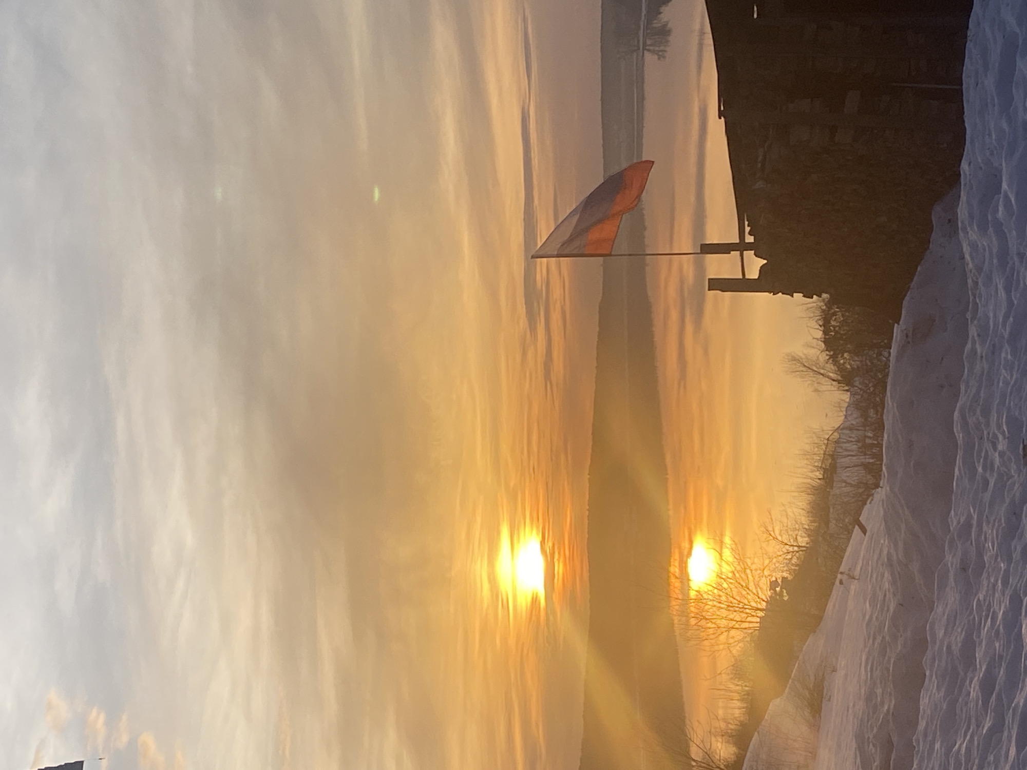 The image depicts a winter landscape in the city of Ольховка, Russia, at either sunrise or sunset. The foreground presents a snow-covered incline with uneven terrain. Sparse, bare deciduous vegetation is visible, particularly on the left. To the lower right, a dark, weathered structure, possibly an outbuilding constructed from stacked logs or stones, is present. A flagpole extends from this structure, flying a Russian flag (horizontal white, blue, and red stripes) oriented to the left. The midground features a wide, calm body of water reflecting the brilliant orange and yellow light. The sun is prominently positioned low on the horizon, slightly left of center, emitting strong rays and creating a distinct reflection on the water's surface. A dark, indistinct landmass forms the opposing bank. The sky above is partially cloudy, displaying warm gradients of orange, yellow, and pale blue. No individuals or specific actions are depicted.