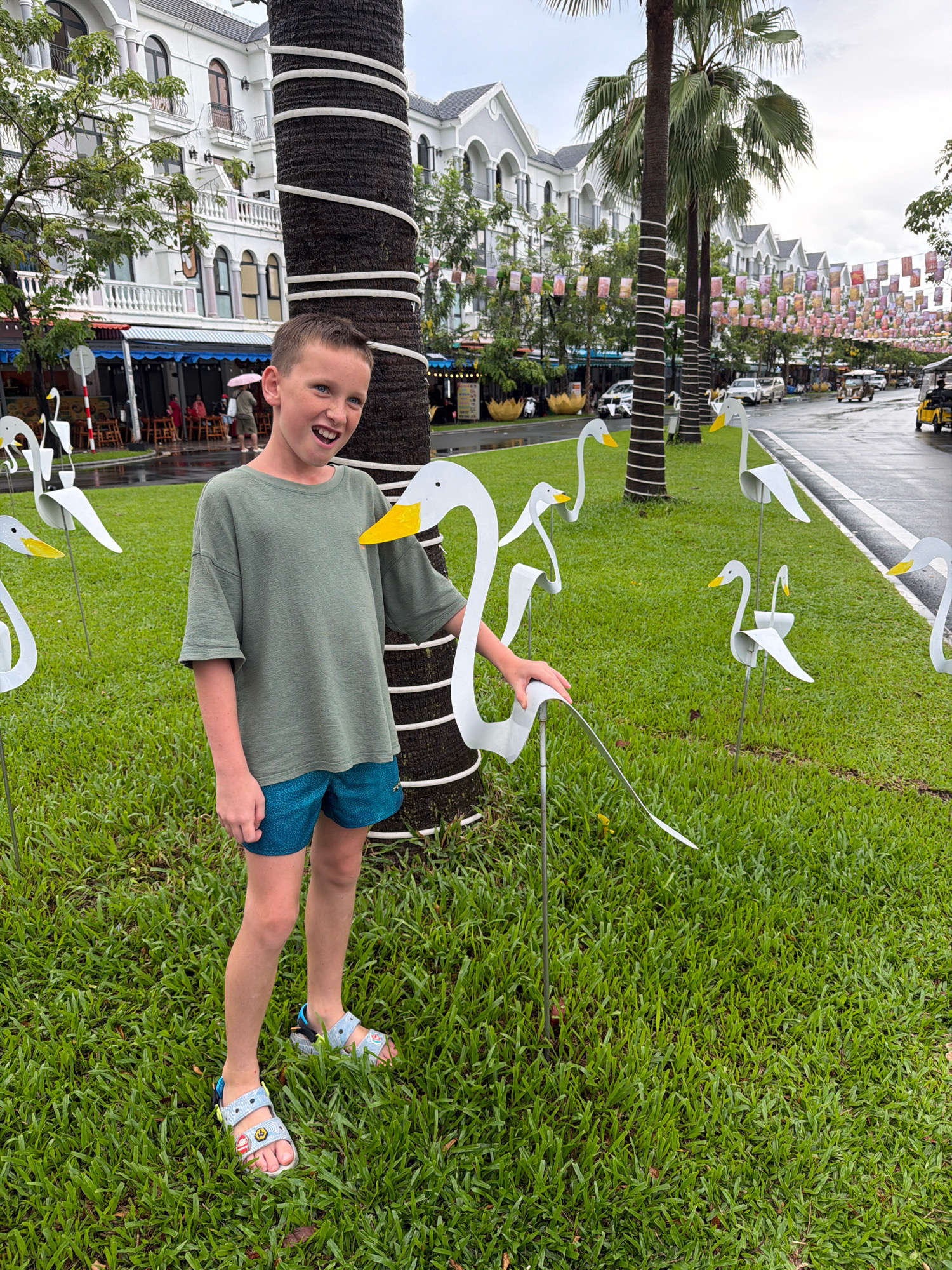 In Phú Quốc, Vietnam, a young boy with light brown hair and blue eyes stands on a grassy lawn next to a large palm tree wrapped in white tape. He is wearing a green t-shirt and blue shorts, with light blue sandals on his feet. The boy is smiling broadly, with his mouth open. In front of him and to his left are several white, abstract sculptures of swans with yellow beaks, some of which are partially obscured by his body. To his right, the lawn continues, lined with more swan sculptures, a paved road with vehicles, and buildings with white facades and arched windows. The sky is overcast, suggesting a cloudy day. The overall impression is a playful outdoor setting decorated with decorative elements.