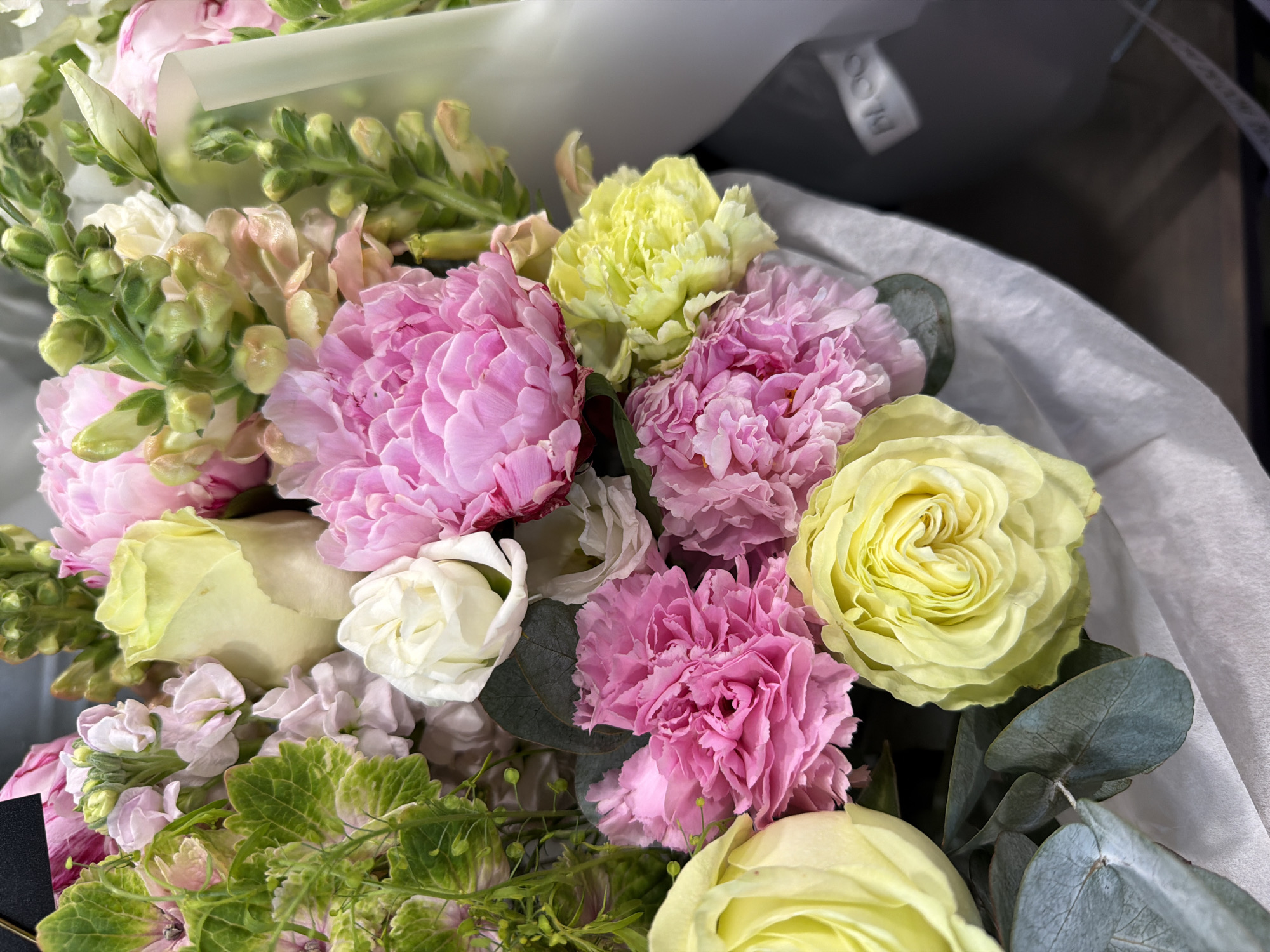 A close-up, overhead shot showcases a vibrant bouquet of mixed flowers. In the foreground, bright yellow roses with full, layered petals are prominent. To their left, a cluster of soft pink carnations with ruffled edges is visible, alongside delicate white roses. Further back, lush pink peonies with densely packed petals add volume to the arrangement. Interspersed among the blooms are spires of pale pink and white snapdragons and sprigs of green foliage, including eucalyptus leaves. The bouquet is loosely wrapped in white paper, with a dark grey container partially visible at the bottom right. A small black tag with white lettering is partially obscured in the upper left corner.