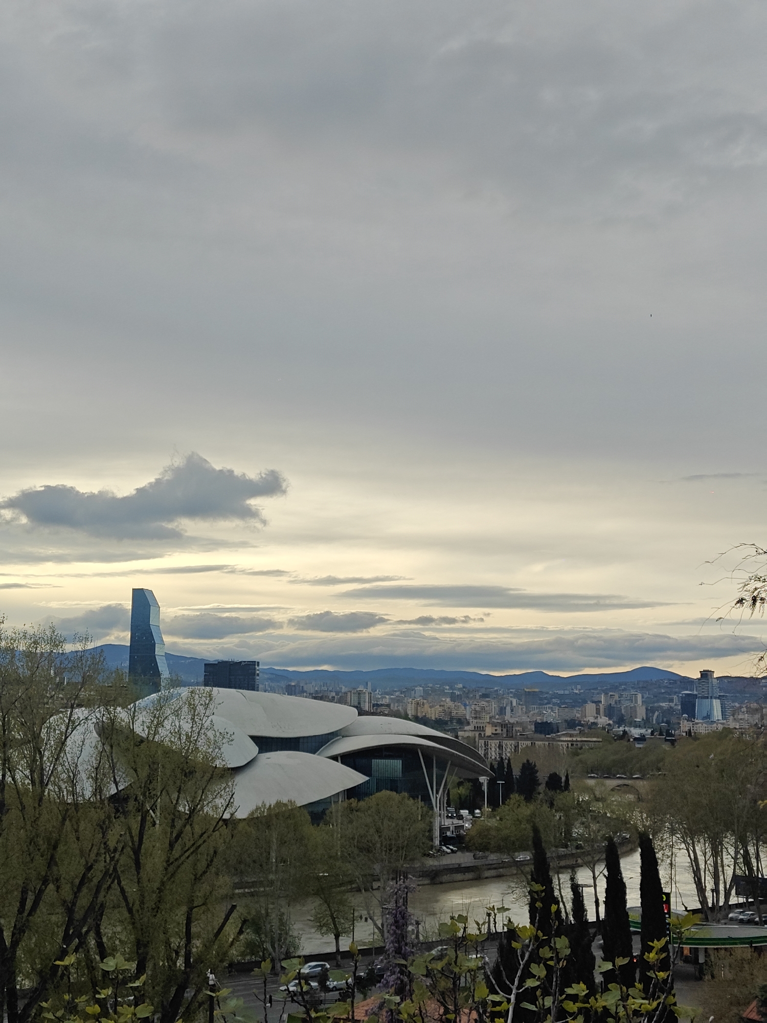 A panoramic view of Tbilisi, Georgia, captures a modern urban landscape under an overcast sky. The scene is dominated by contemporary architecture and natural elements.
In the midground, the Kura (Mtkvari) River winds through the city, flanked by roads and trees. Rolling hills and mountains form the distant backdrop, framing the cityscape.
The sky is cloudy, presenting various shades of grey, with hints of soft, golden light filtering through, suggesting either early morning or late afternoon. A notable cloud formation to the left of a tall skyscraper strikingly resembles an animal, possibly a dragon or a dog.
Two prominent buildings stand out: a tall, angular glass skyscraper with a distinctive faceted design, and a sprawling complex below it featuring multiple white, shell-like or mushroom-domed roofs, identifiable as the Public Service Hall. Another dark, modern building is situated near the skyscraper.
The foreground is filled with lush green trees, some displaying fresh spring foliage, partially obscuring the view of the riverbanks where several cars and a green-canopied gas station are visible. A bridge can be seen spanning the river further in the distance.
No specific human activity or visible text is discernible in the image, which presents a serene, atmospheric vista of the city.