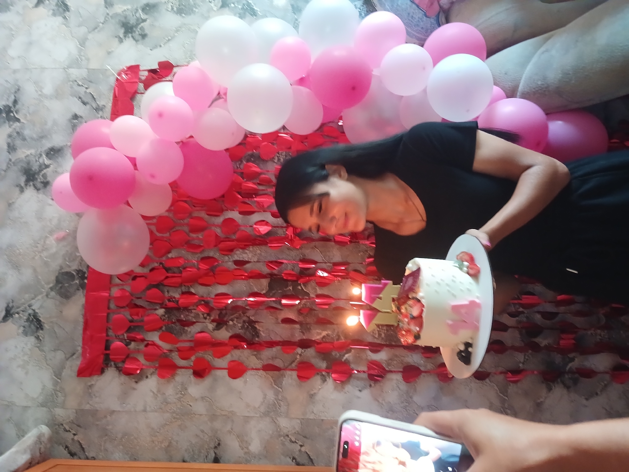 A young woman in a black dress stands in a room decorated with pink and white balloons and red heart garlands. She is holding a white cake topped with strawberries and has the number "14" candles on it. The woman is looking down at the cake with a smile. The background shows a textured wallpaper and a couch. The image appears to be taken indoors, likely in a residential setting. There is no information about the city of Chik, Russia.