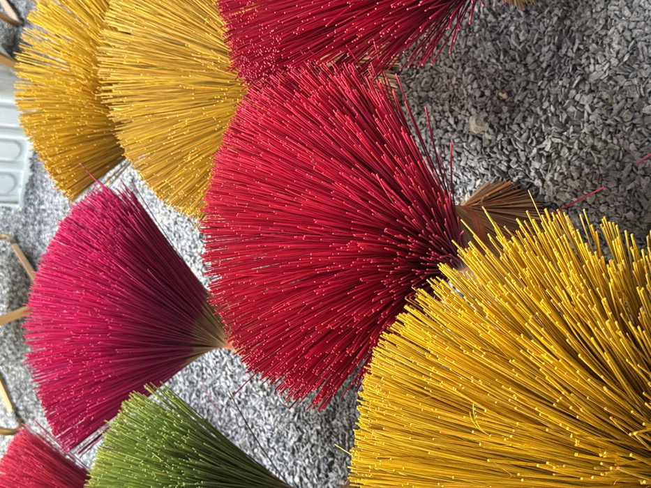 Colorful bundles of incense sticks, predominately red and yellow, arranged on a textured surface. The sticks are fanned out, forming semi-circular shapes. Some bundles appear pink and green. The background is a coarse, gray material resembling gravel. The image is taken from a high angle, providing a close-up view of the incense bundles and their arrangement.