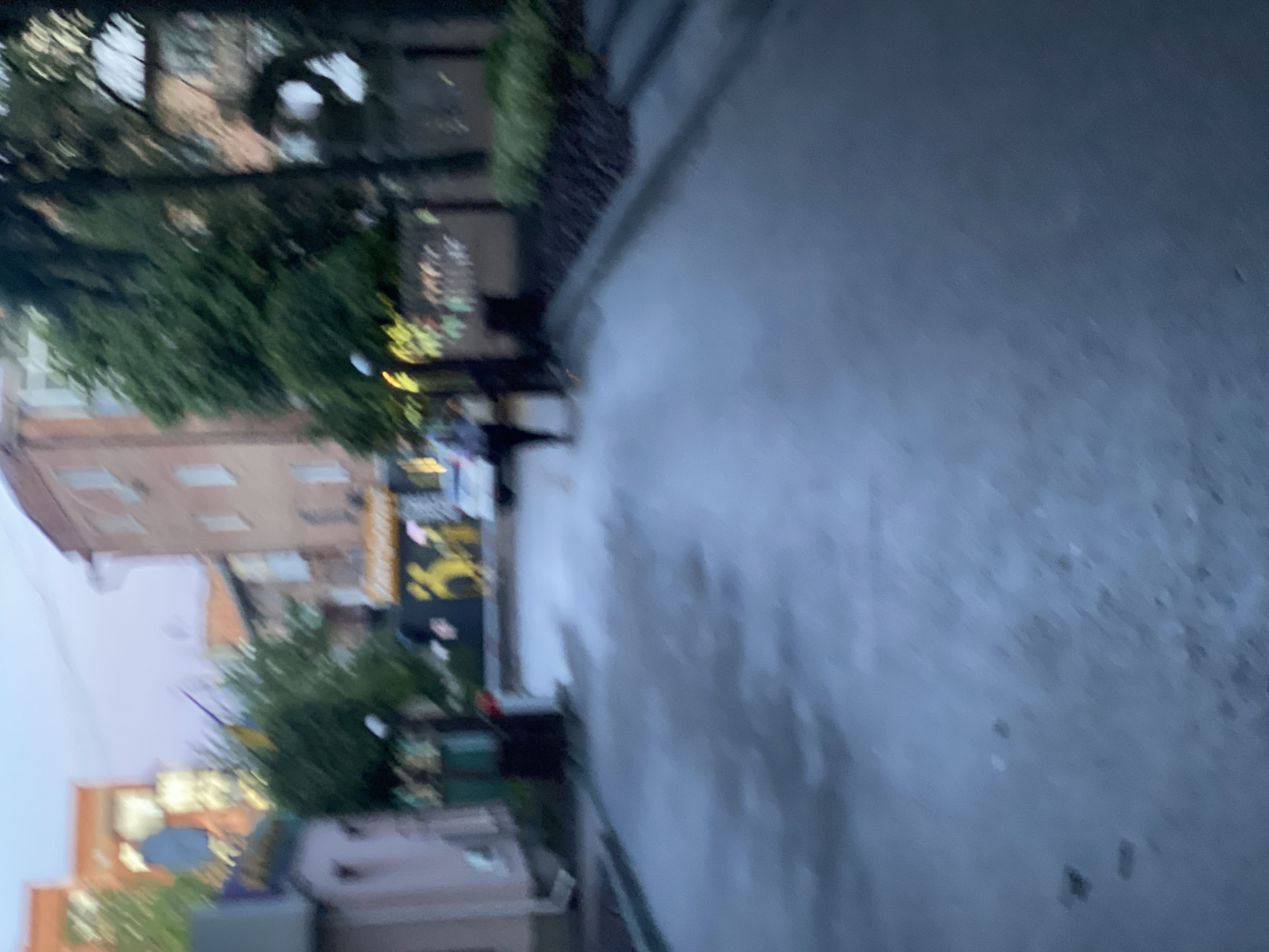 The image shows a street scene in the city of Poltava, Ukraine. The view is partially obscured by a curb or wall that takes up a large portion of the right side of the frame. On the left side, buildings can be seen, along with trees and street foliage. There are signs and lighting indicating commercial activity or a public area. A person or persons may be present, but the image quality and orientation makes it difficult to discern distinct individuals or their specific actions. The overall environment appears urban.