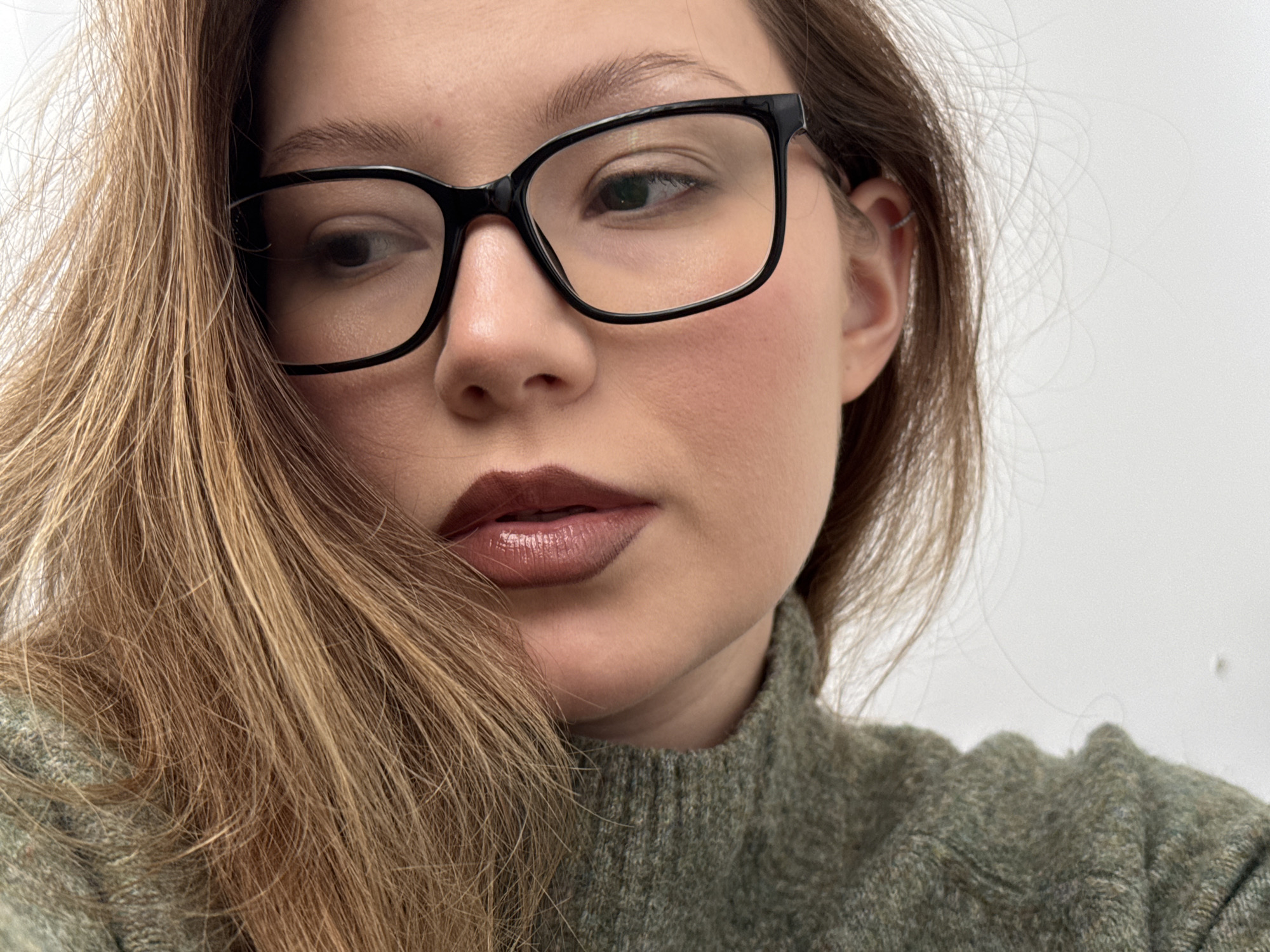 A close-up shot depicts a woman with light skin wearing black-framed glasses and dark lipstick. She is looking to the left with a contemplative expression. The woman has long, light brown hair and is wearing a green turtleneck sweater. The background is a plain white wall.