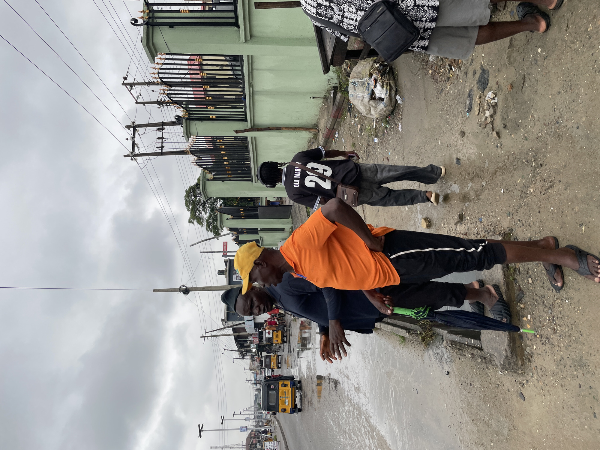 The image shows a street scene in Ado, Nigeria, on a cloudy day. The road is flooded with water reflecting the overcast sky. Several yellow-colored auto rickshaws, locally known as "Keke Napep," are visible in the distance. Three men are walking along the edge of the flooded street: one in an orange shirt and yellow cap, one in a black shirt, and another further back wearing a black jersey with the number 29. A green building with a black metal fence is present on the right side of the image. Power lines and utility poles are seen overhead. The environment appears urban and somewhat congested. Litter is visible along the side of the road.