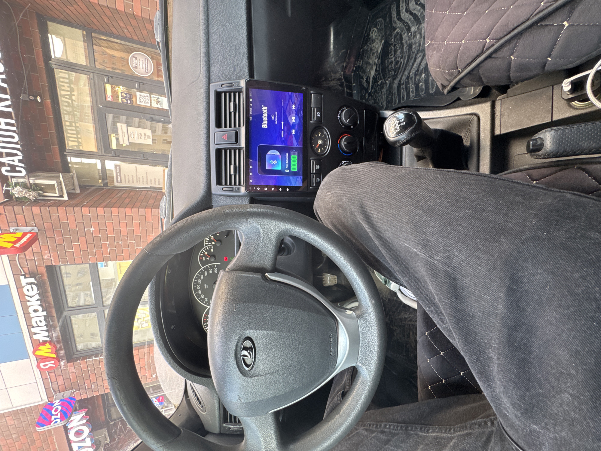 A photograph taken from inside a Lada vehicle, shows a steering wheel and the dashboard, with a person sitting in the driver's seat. The car's infotainment screen displays a Bluetooth connection interface. Outside the window, signs reading "Я Маркет" and "САЛОН" (salon) are visible, likely indicating a store or beauty salon located in Perm, Russia. A building exterior with brick facade is also apparent. The gear shift lever is also shown.