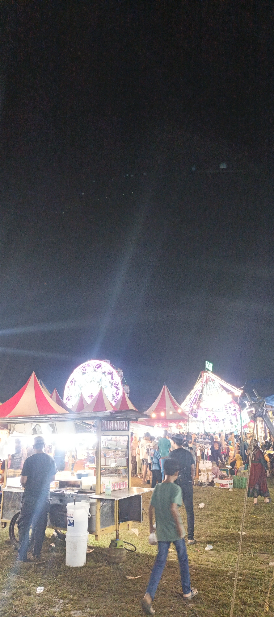 The image depicts a night scene at an outdoor fair or market in Banding Agung, Indonesia. The sky is dark, with a few scattered lights and what appear to be distant lights from the city or the fair itself. In the foreground, a young boy in a green t-shirt and blue jeans walks towards the right of the frame, carrying a plastic bag. Behind him, a man in a dark t-shirt and cap stands facing away from the camera, near a food cart with a "MARTABAK" sign. The food cart appears to be lit and stocked with items. To the left of the food cart, a large white container is visible.
Further into the background, several tents or stalls with red and white striped awnings are set up. Dominating the middle ground and background is a large, brightly lit Ferris wheel, which is circular and illuminated with white and pink lights. Another large, cone-shaped tent, also illuminated with colorful lights, is visible to the right of the Ferris wheel, topped with a green sign. Numerous people are milling about in the background, suggesting a lively atmosphere. The ground is covered with grass and scattered litter. The overall impression is that of a bustling night market or carnival.